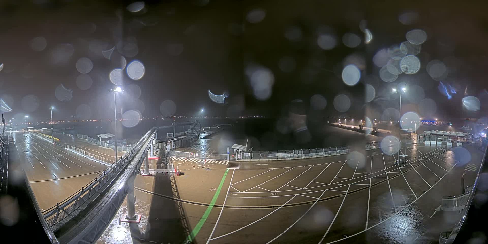 A rain-soaked, nighttime view of an empty port or ferry terminal with marked parking areas and a gangway, viewed through a rain-covered window.