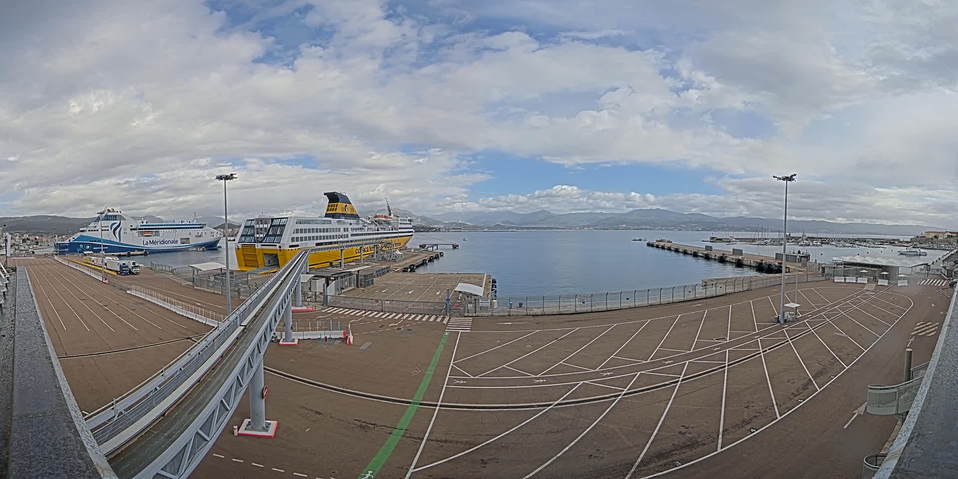 Two large ferries are docked at a port under a partly cloudy sky, with a paved area and a walkway leading to them.
