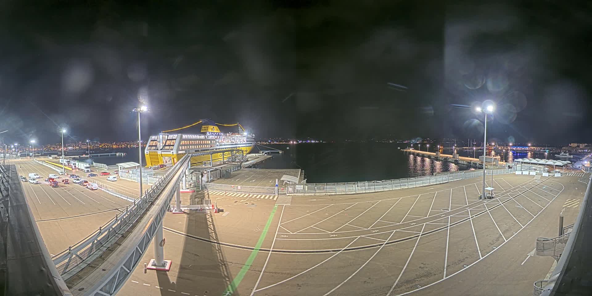 At night, a large yellow and blue ferry is docked at a port with cars parked nearby under mostly cloudy skies.