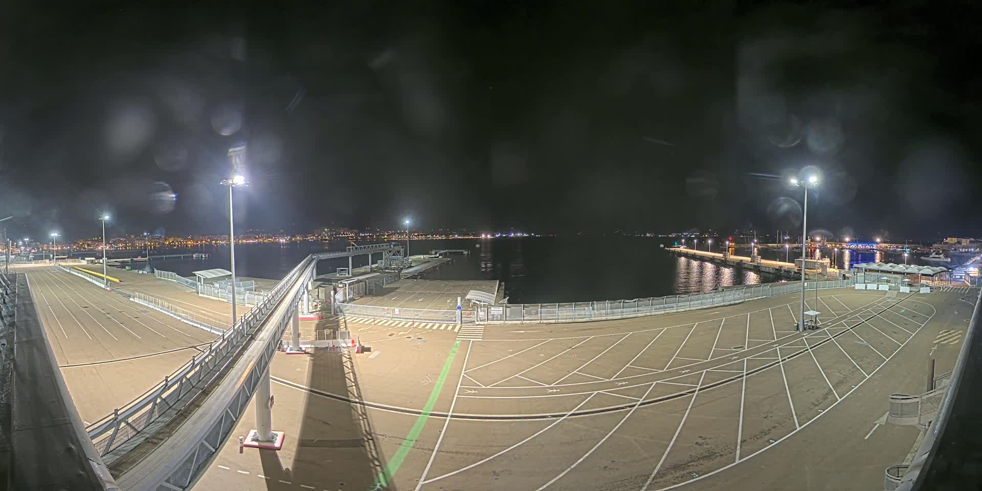 An empty, illuminated port at night, viewed from above, with a calm body of water in the background under a dark, slightly misty sky.