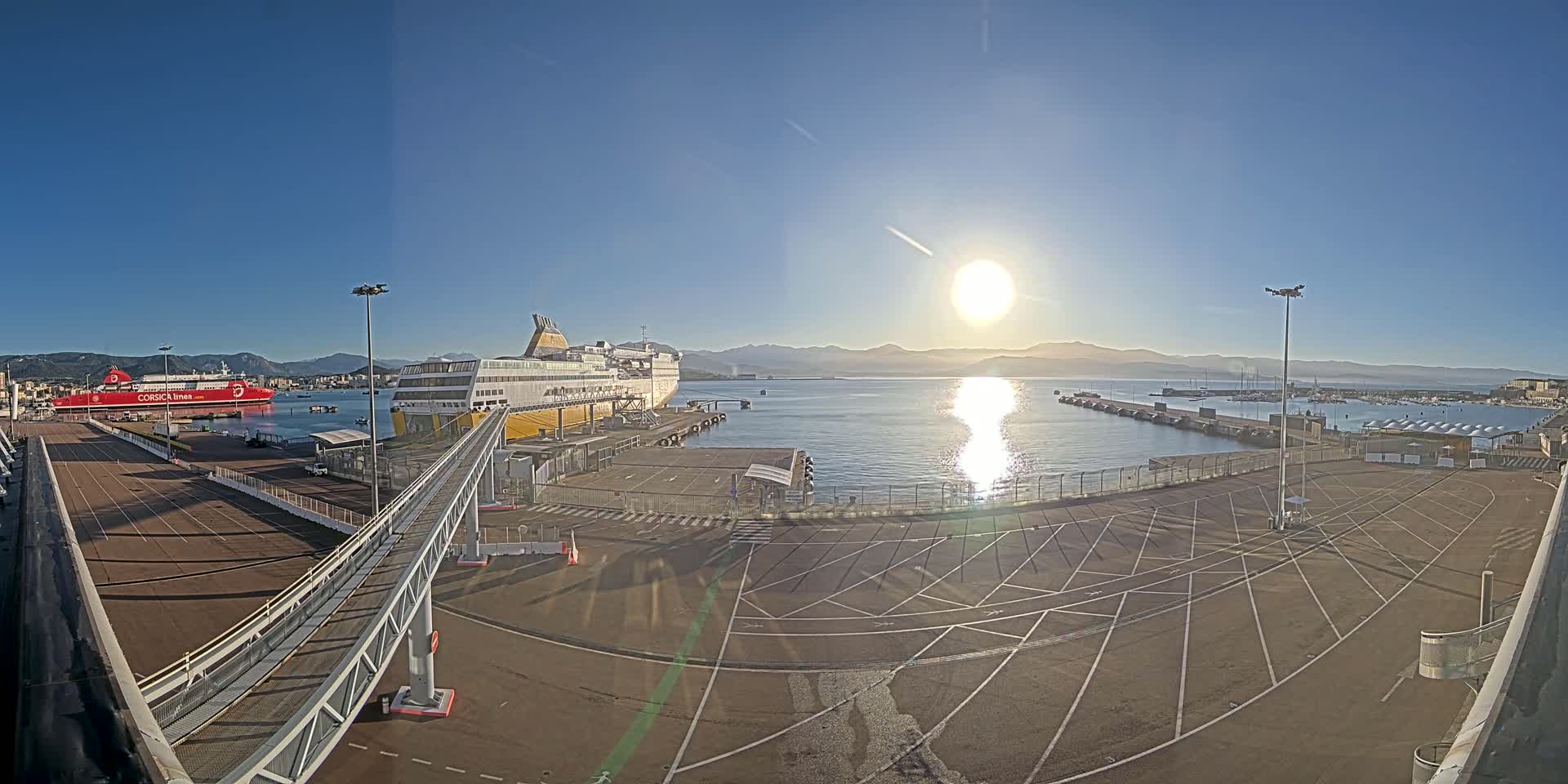 A sunny day at a port shows two large ferries docked, with a walkway leading to one, and an empty parking lot in the foreground.