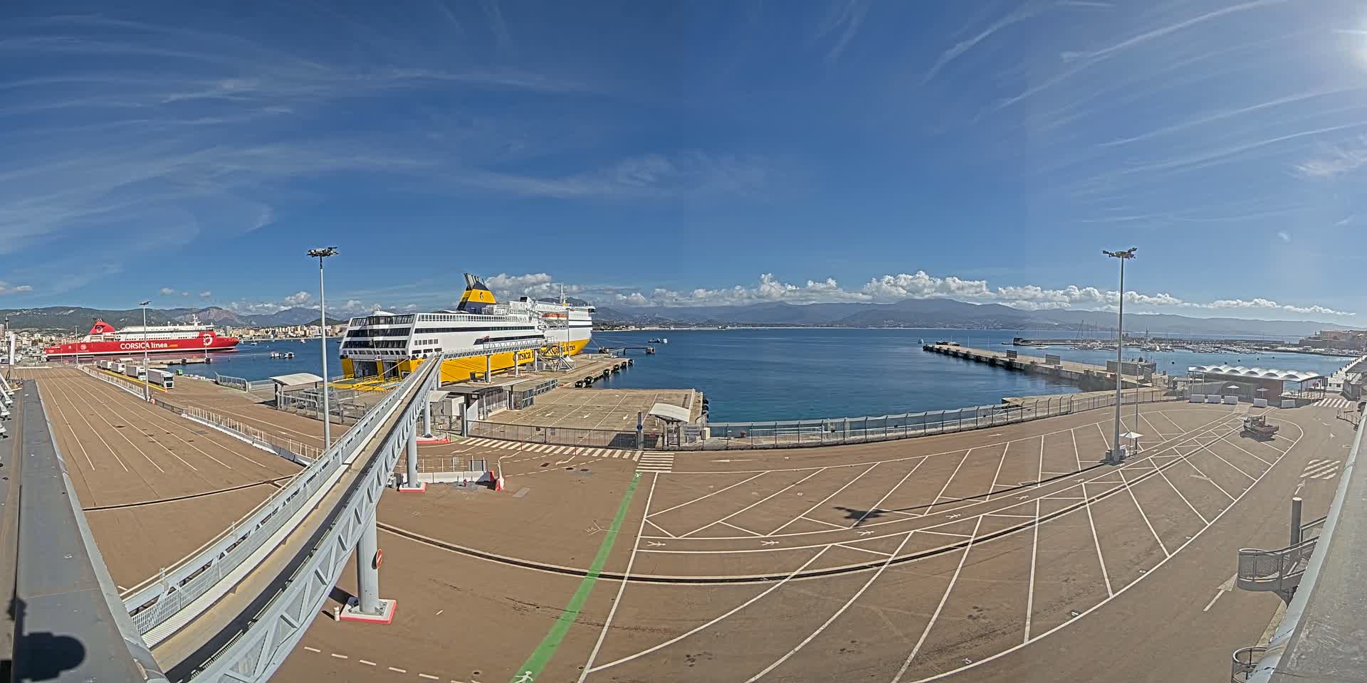 A ferry terminal with two docked ferries under a mostly sunny sky, mountains in the distance, and a large empty parking lot.