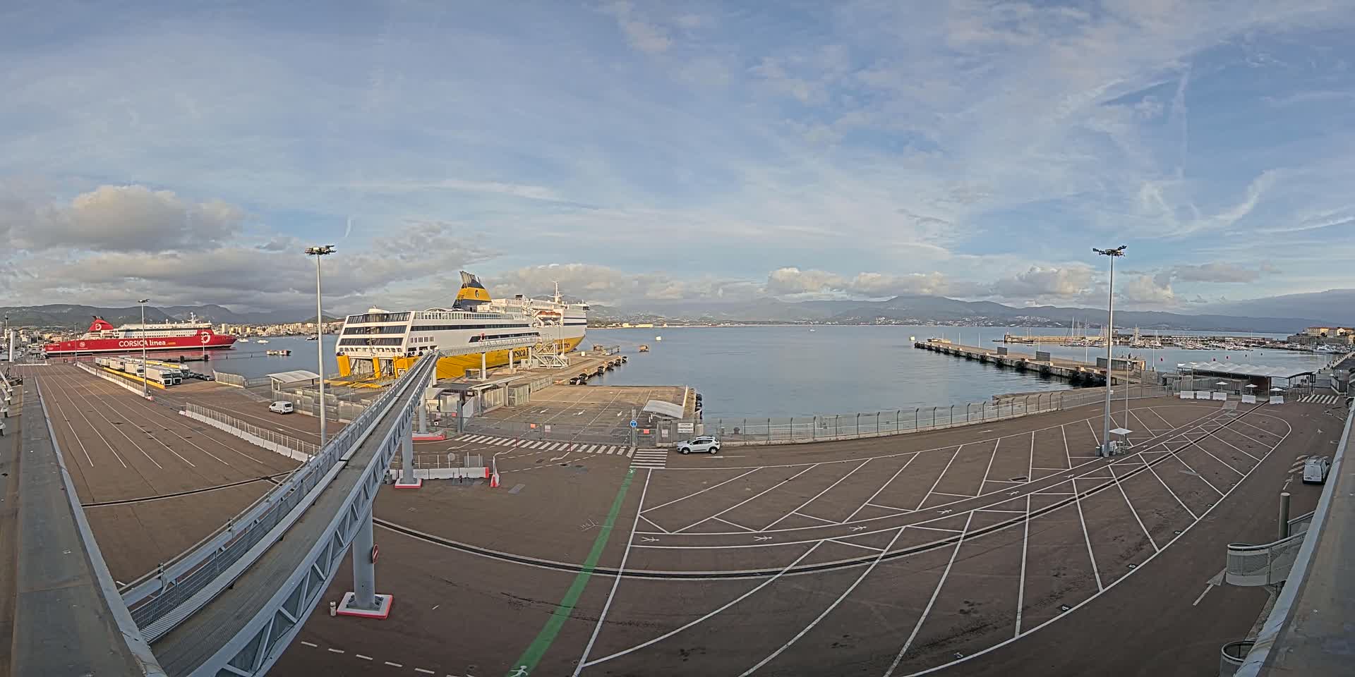 A large port area with two docked ferries, a mostly empty parking lot, and a gangway leading to one of the ships under a partly cloudy sky.
