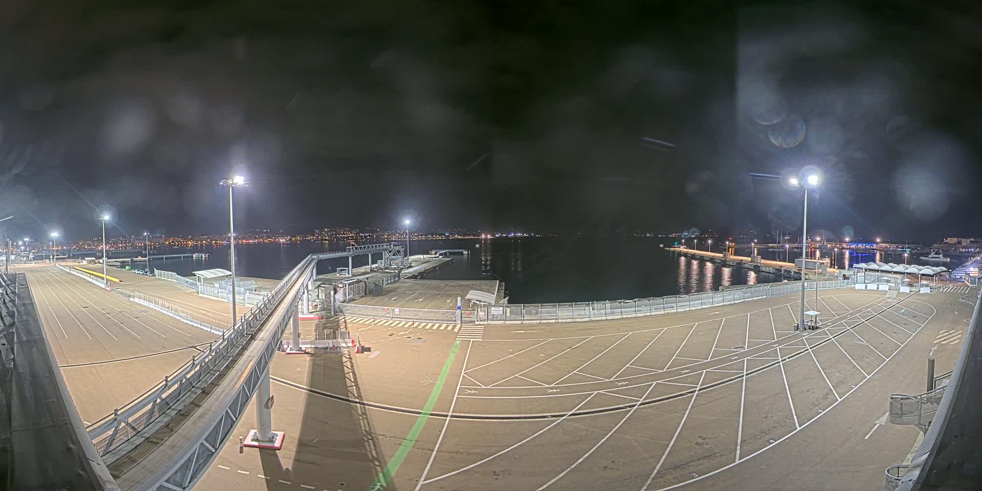 An empty, nighttime ferry terminal with marked parking spaces overlooks a calm body of water and a distant city skyline under a dark, cloudy sky.