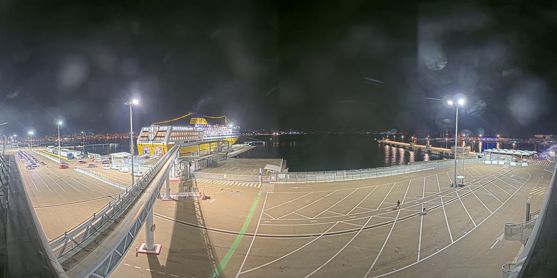 A large ferry is docked at a port at night under a slightly overcast sky, with cars parked in a lot leading to a gangway connecting to the ship.