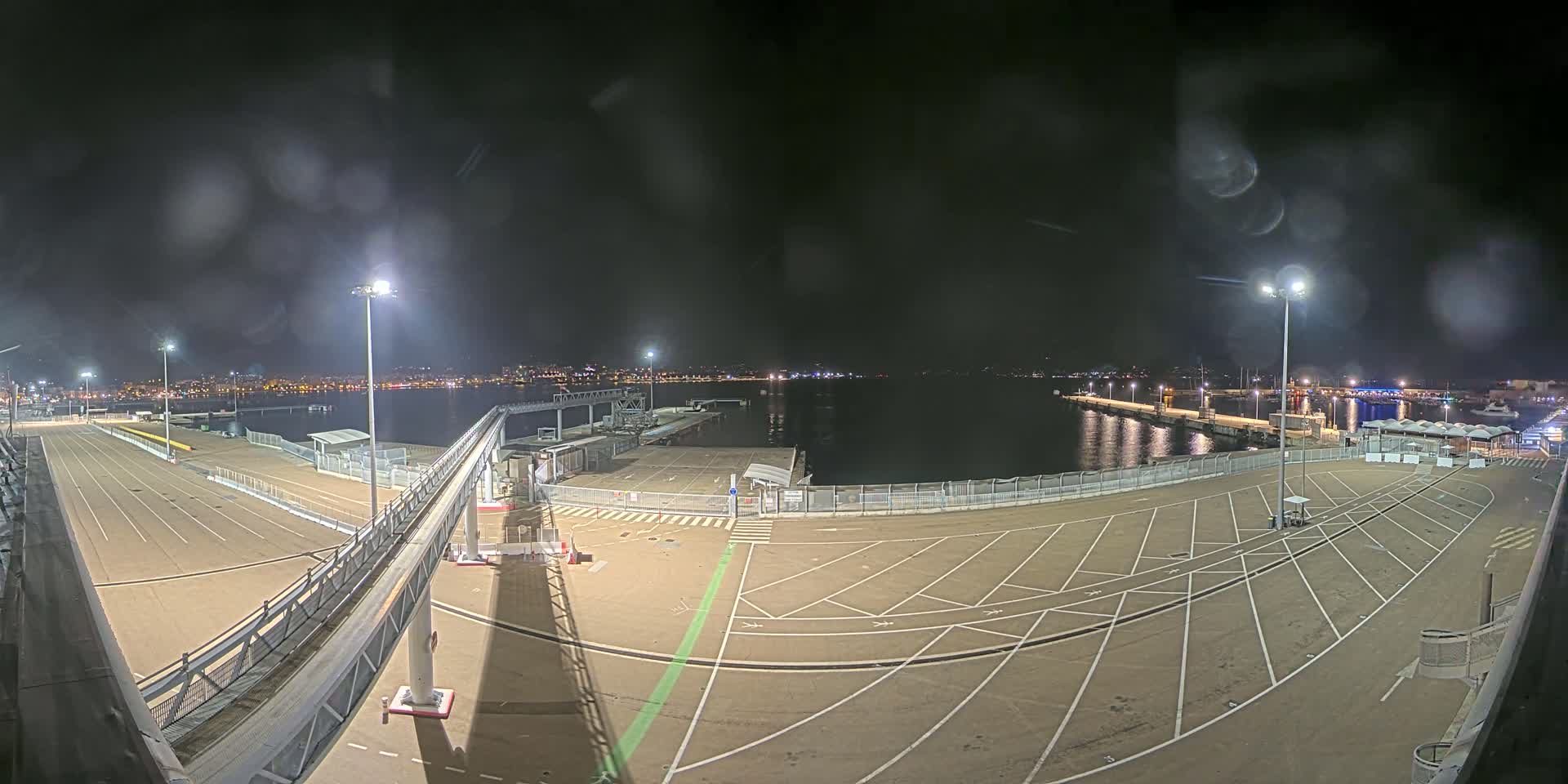 A nighttime view of an empty port area with a gangway, parking spaces, and a body of water showing a distant city skyline under a slightly misty sky.
