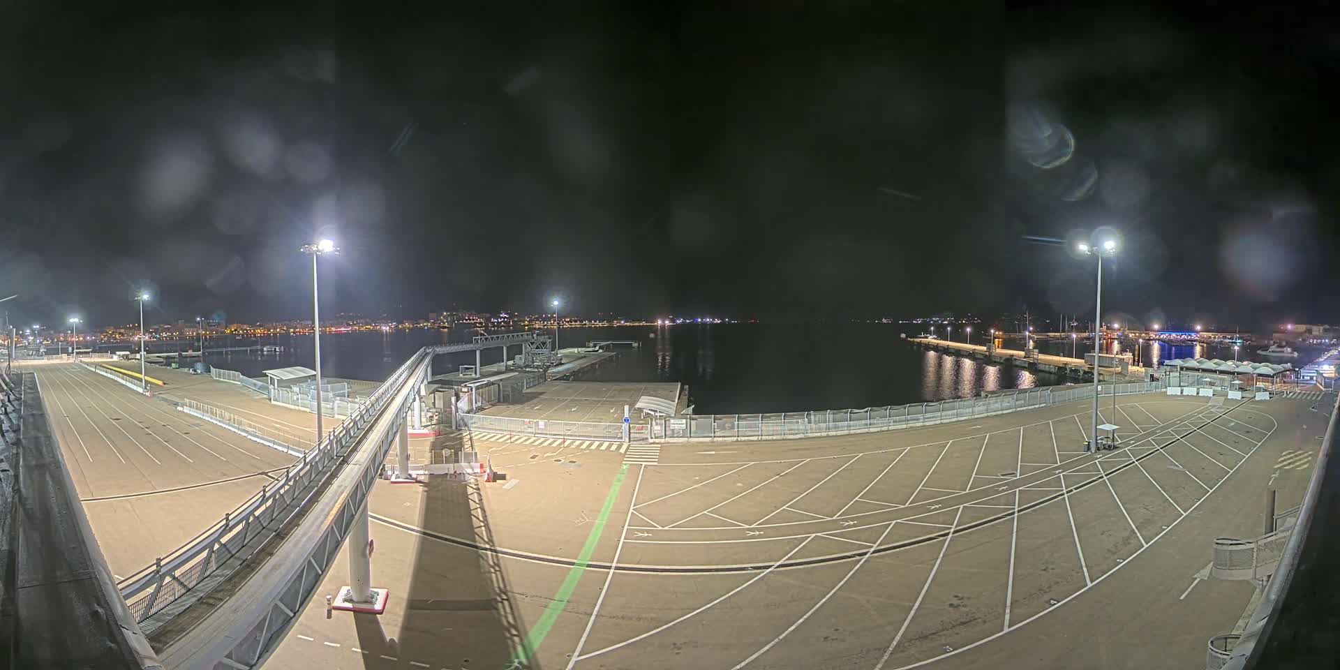 A nighttime view of a mostly empty port area with a gangway, parking lot, and calm water in the background under a slightly hazy sky.