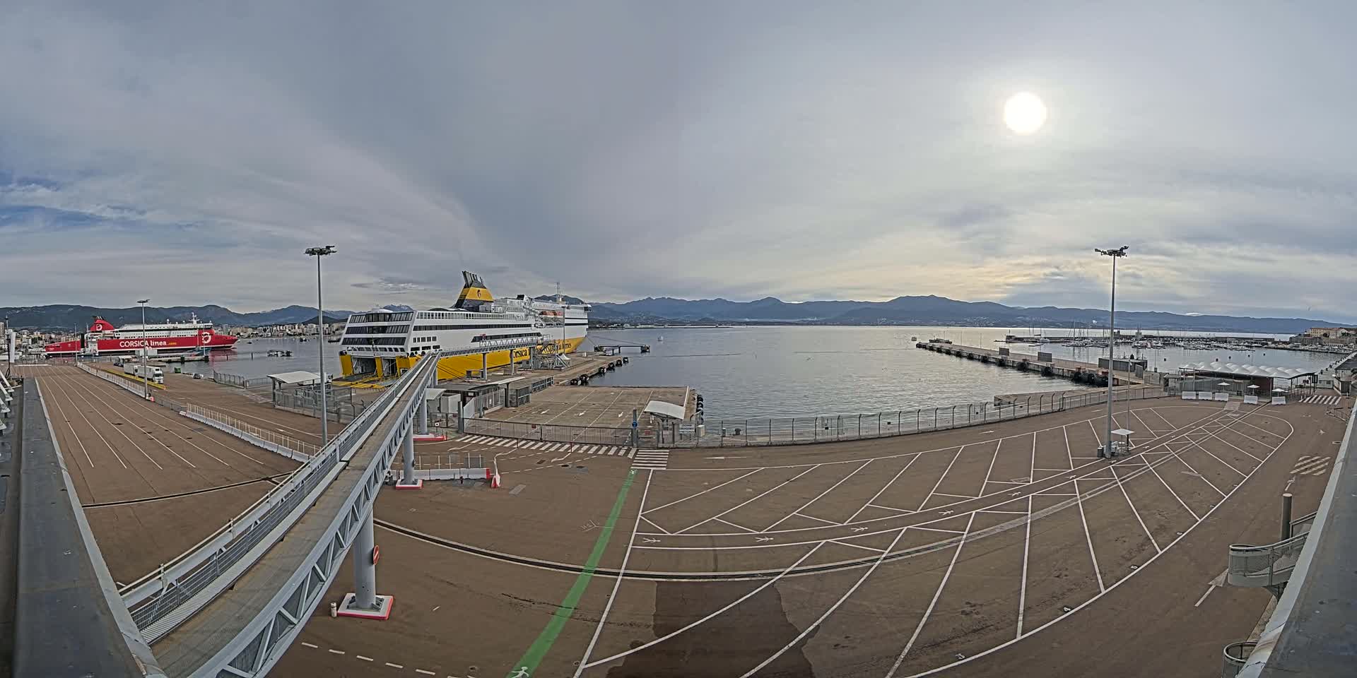 A ferry terminal with two large ferries docked, under a mostly cloudy sky with the sun visible.