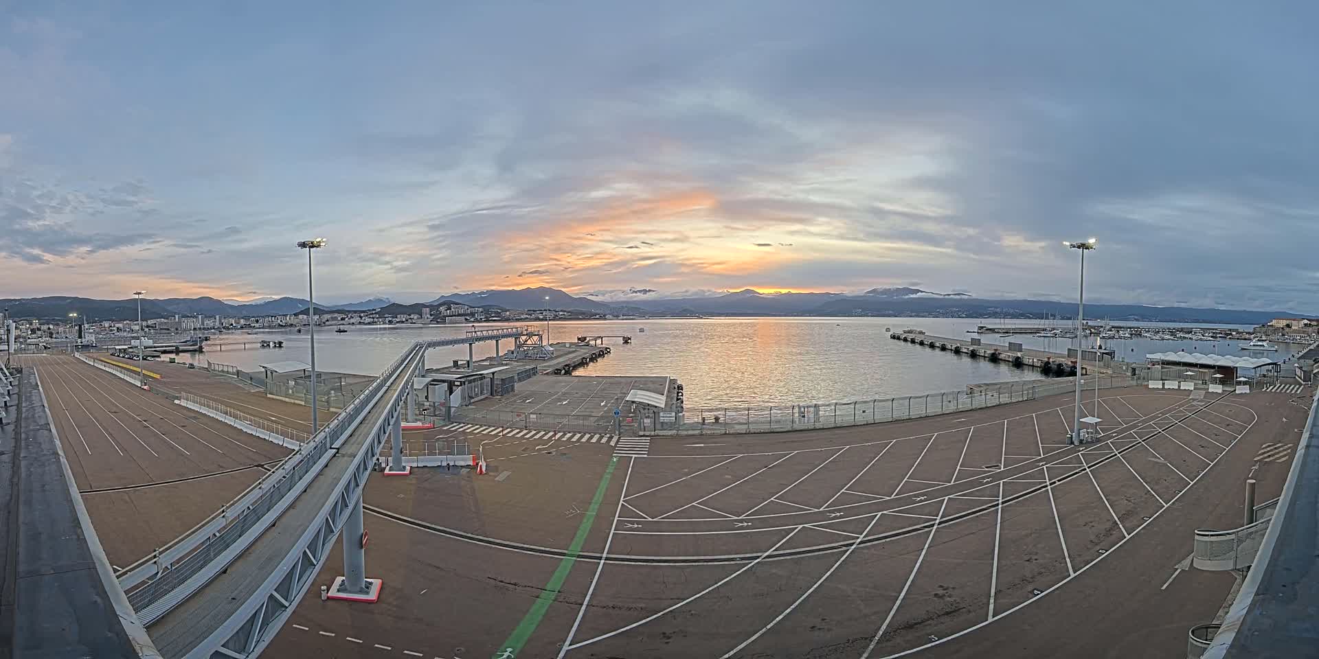 An empty port area with a walkway and parking lot overlooks a calm body of water at sunset, with mountains visible in the distance under a partly cloudy sky.