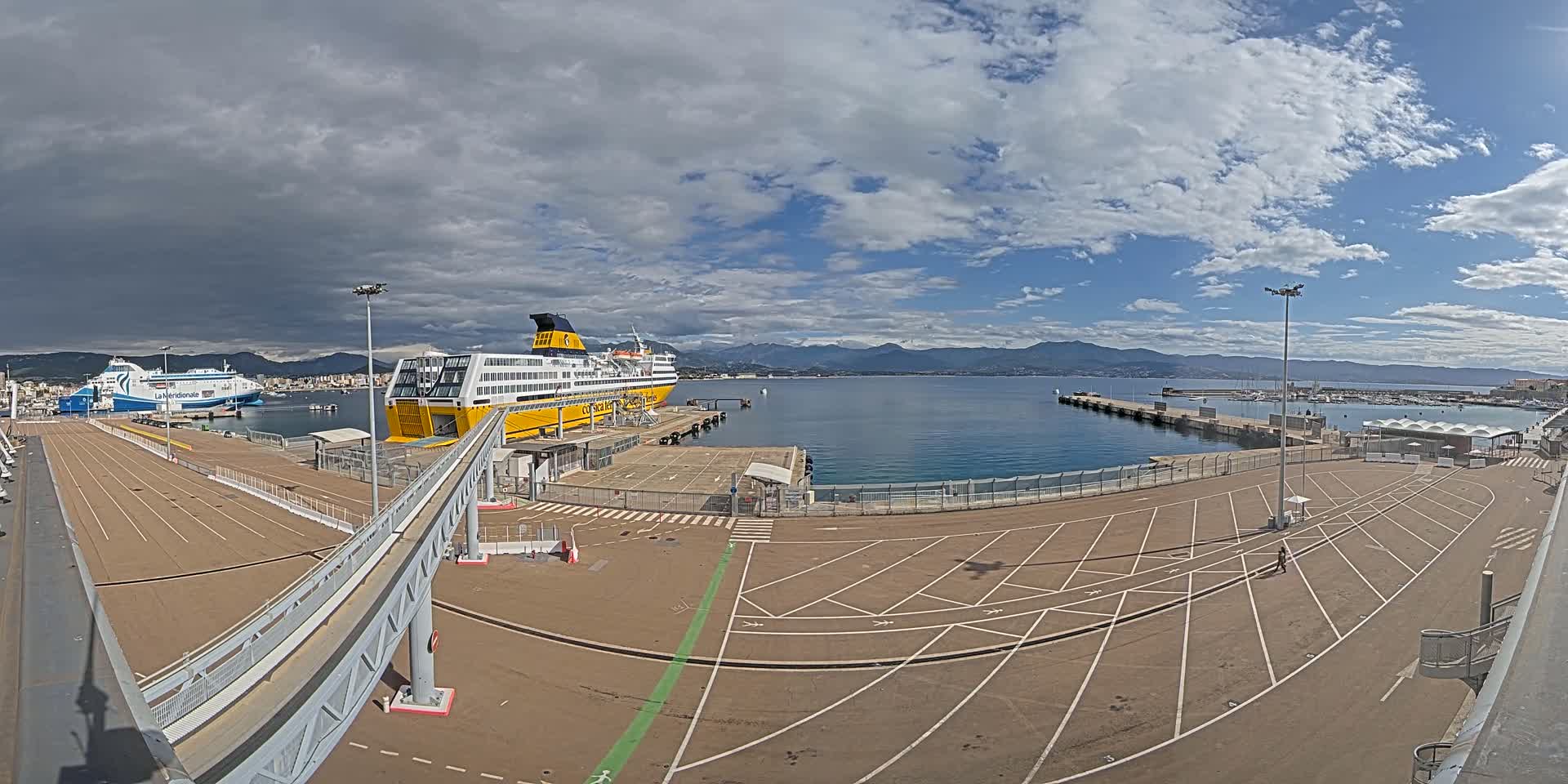 A ferry terminal with two large ferries docked, under a partly cloudy sky, overlooks a calm body of water with mountains in the distance.
