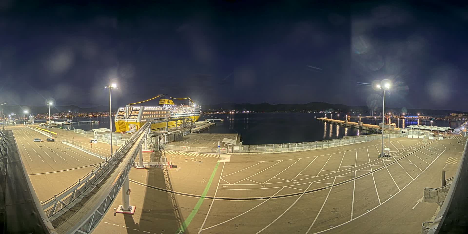 A large yellow ferry is docked at a port at night under a clear, dark sky, with empty parking spaces surrounding it.