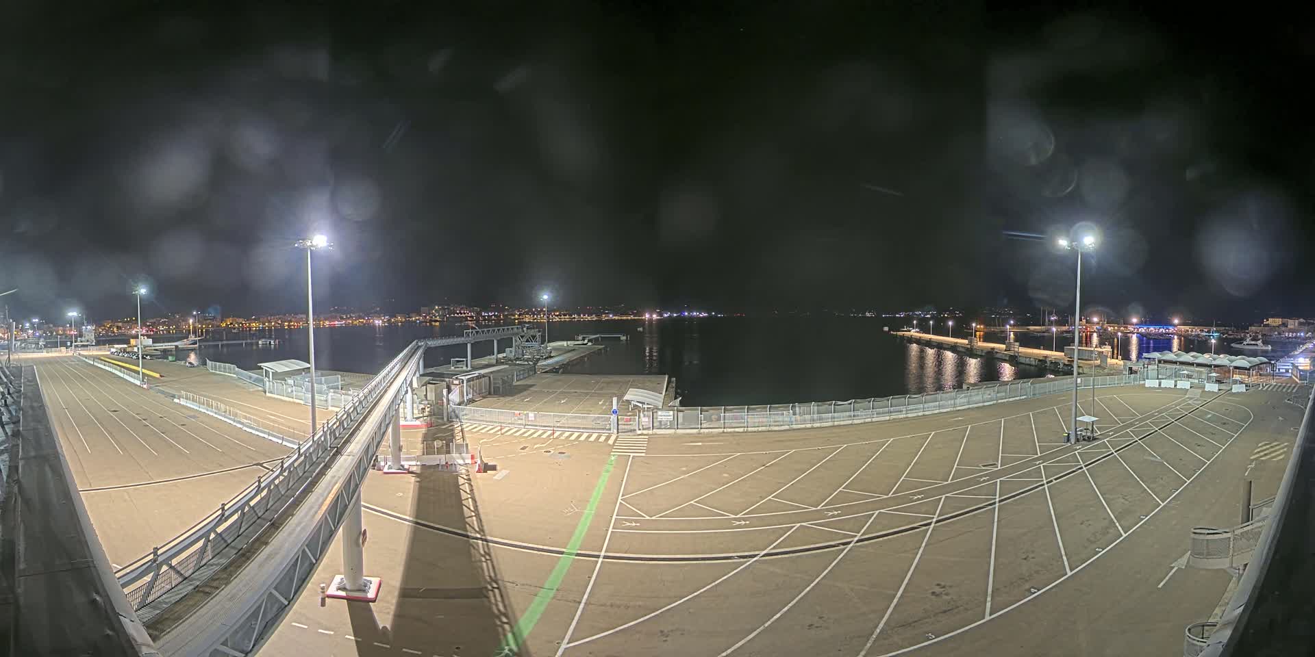 A nighttime view of an empty port area with a passenger gangway,  lit by several bright lamps, under a slightly hazy sky.