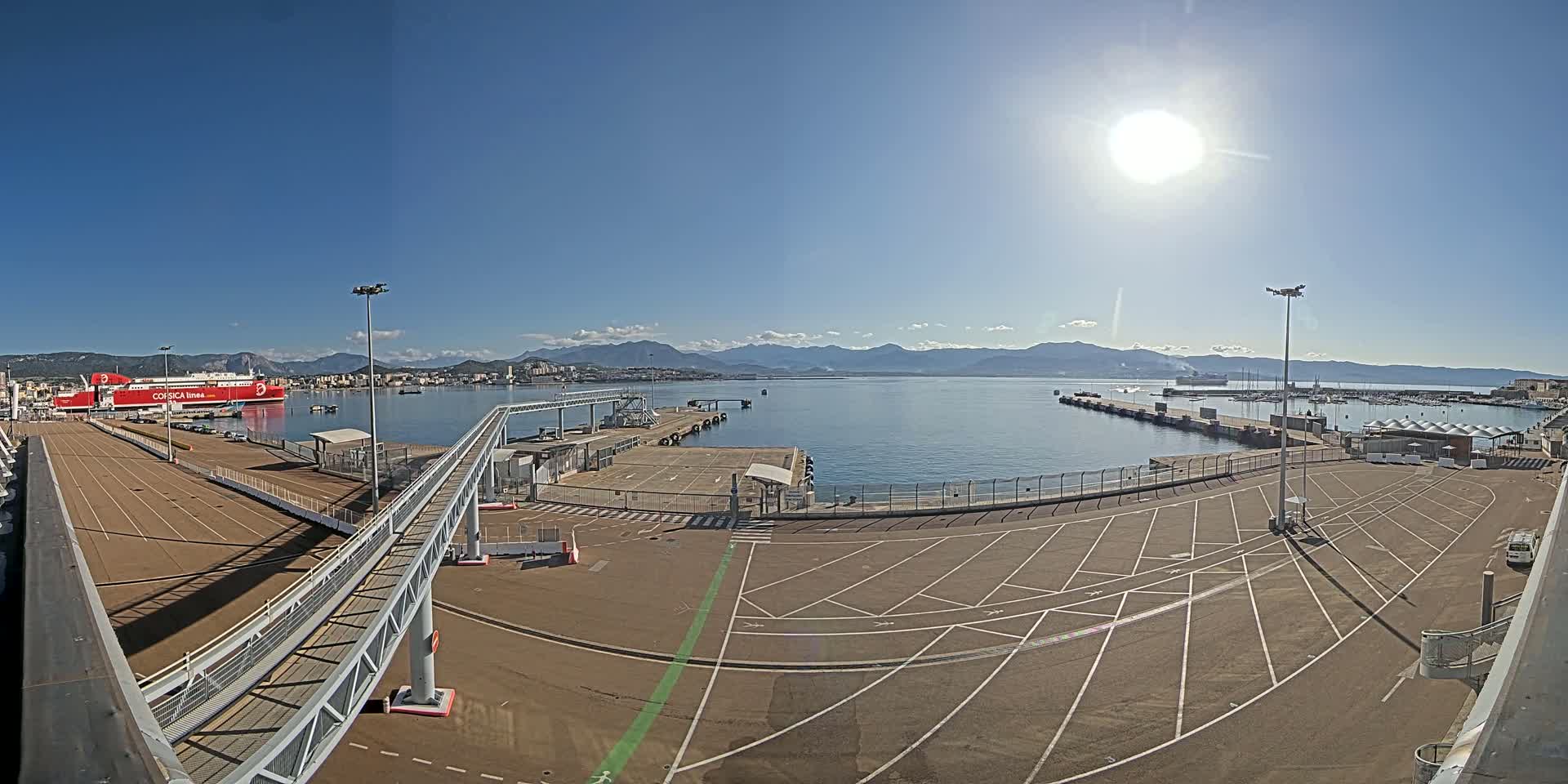 A sunny day at a port, showing a large red ferry docked, empty parking lots, and a walkway leading to a pier, with mountains visible in the distance across calm water.