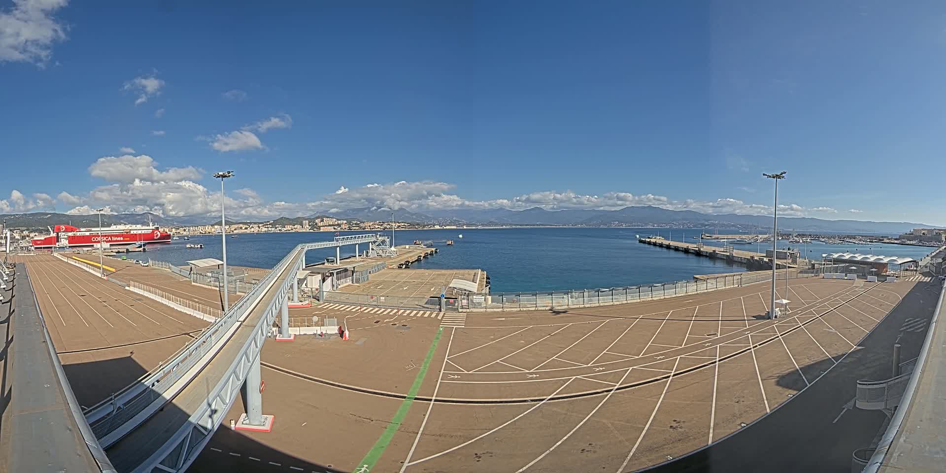 A large, empty port area with a docked red ferry, a walkway leading to it, and a clear, sunny day with distant mountains visible across the calm water.