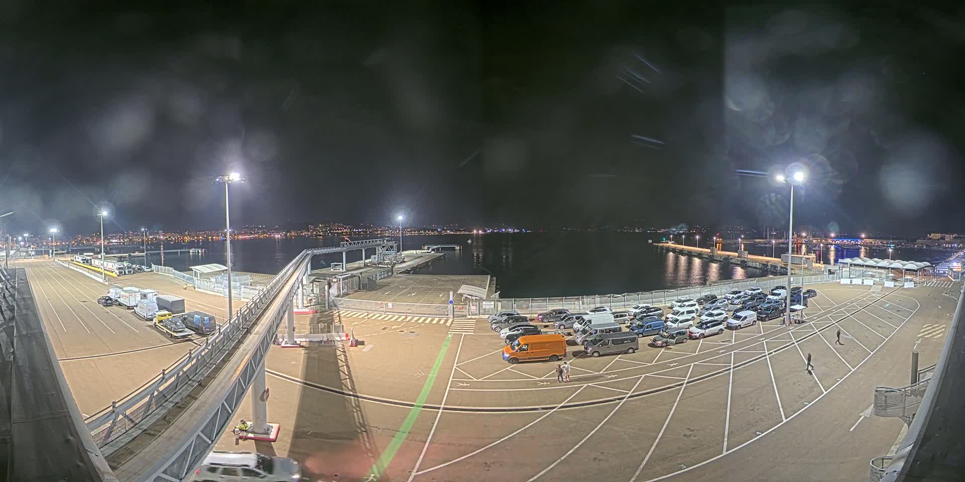 A nighttime view of a ferry terminal with cars parked, a gangway leading to a ship, and a body of water in the background under slightly hazy conditions.
