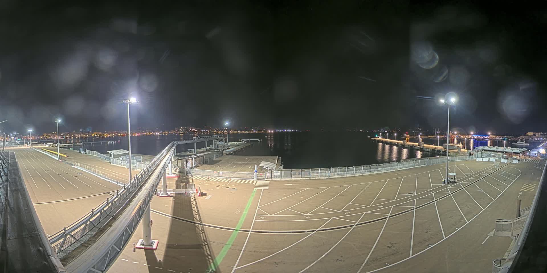 A nighttime view of an empty port area with a gangway, parking spaces, and a calm body of water in the background, slightly obscured by rain or moisture on the camera lens.