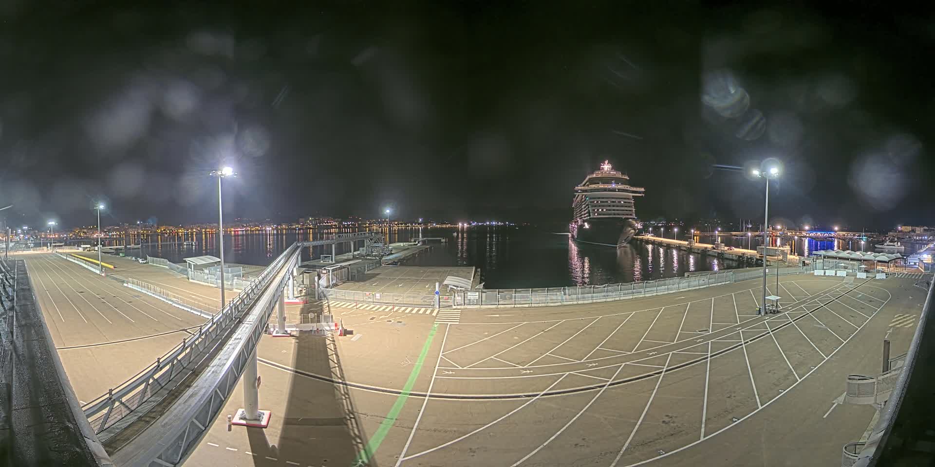 A large cruise ship is docked at a port at night in slightly rainy conditions, with empty parking lots and a gangway visible.