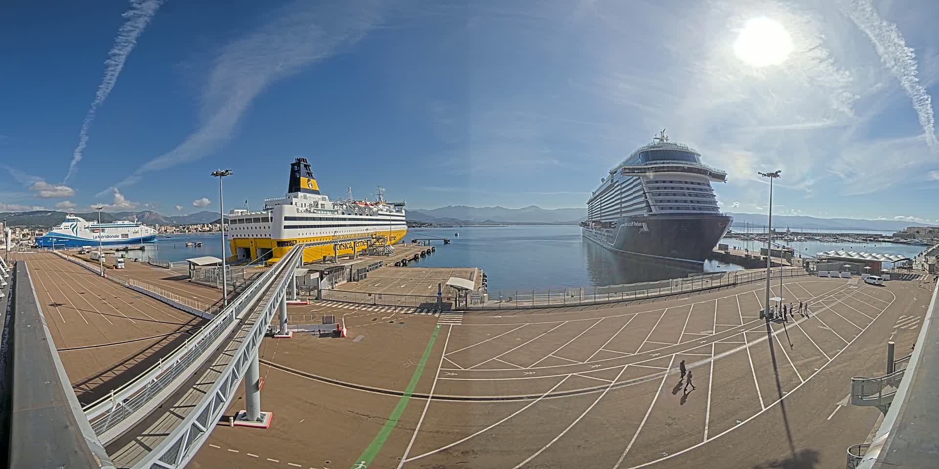 A large cruise ship and two ferries are docked at a port on a sunny day with clear blue skies and mountains in the distance.