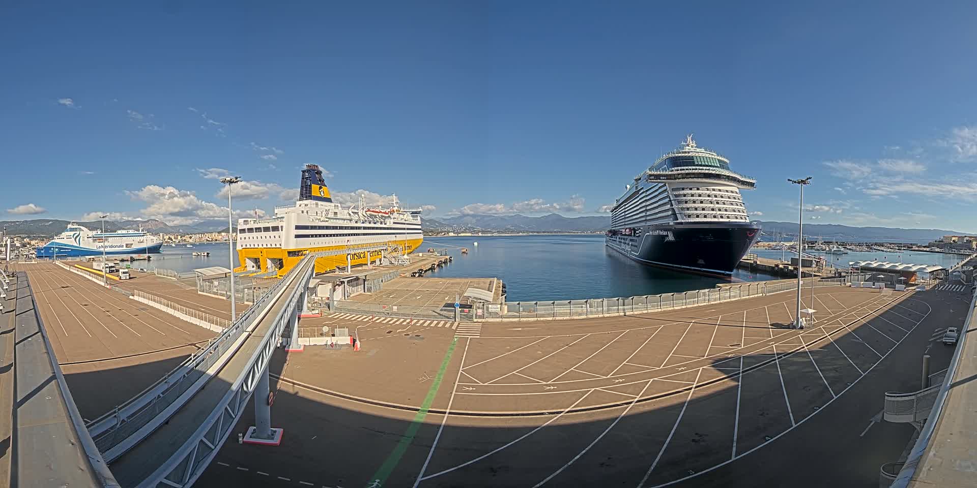 Two large ships are docked at a port on a sunny day with clear skies and mountains in the distance.