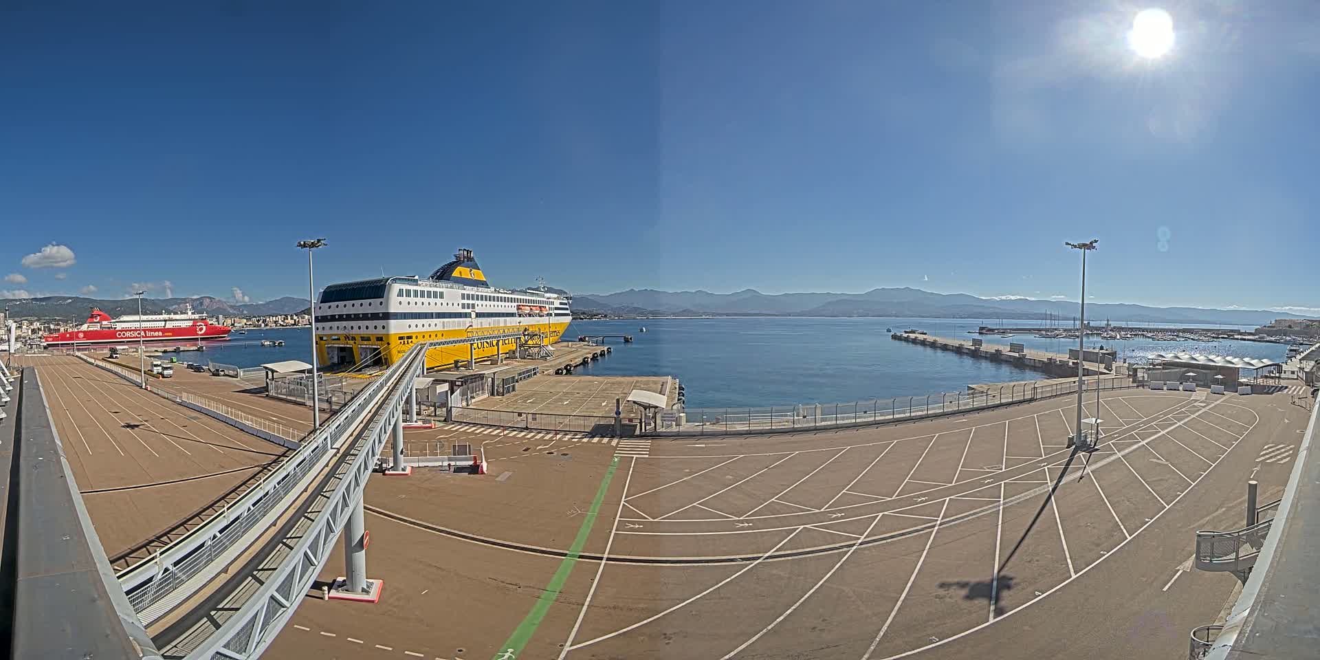 A large yellow and white ferry is docked at a port on a sunny day, with another red ferry visible in the background and mountains in the distance.