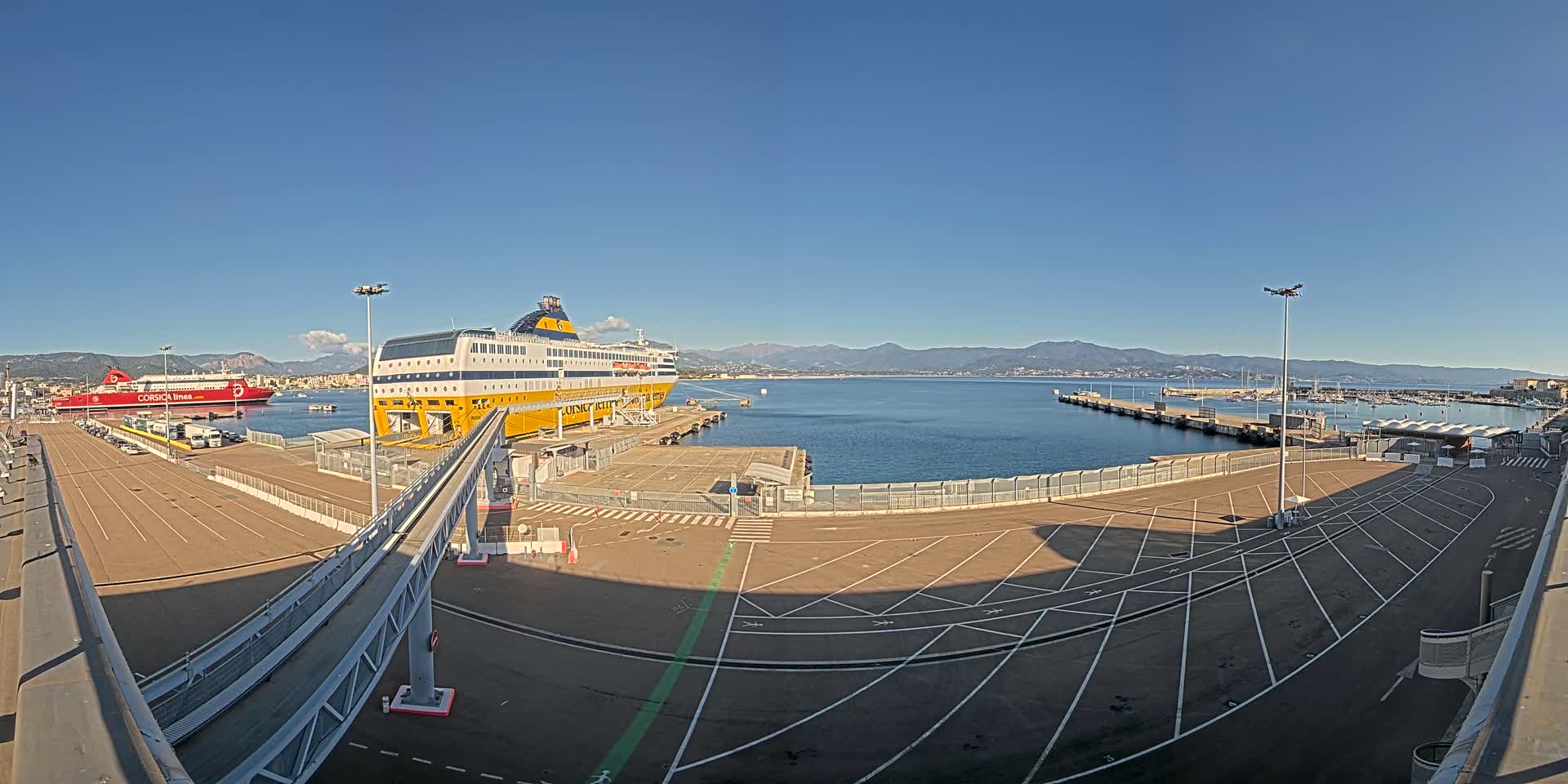 A large yellow and blue ferry is docked at a port on a sunny day, with another red ferry visible in the distance and mountains in the background.