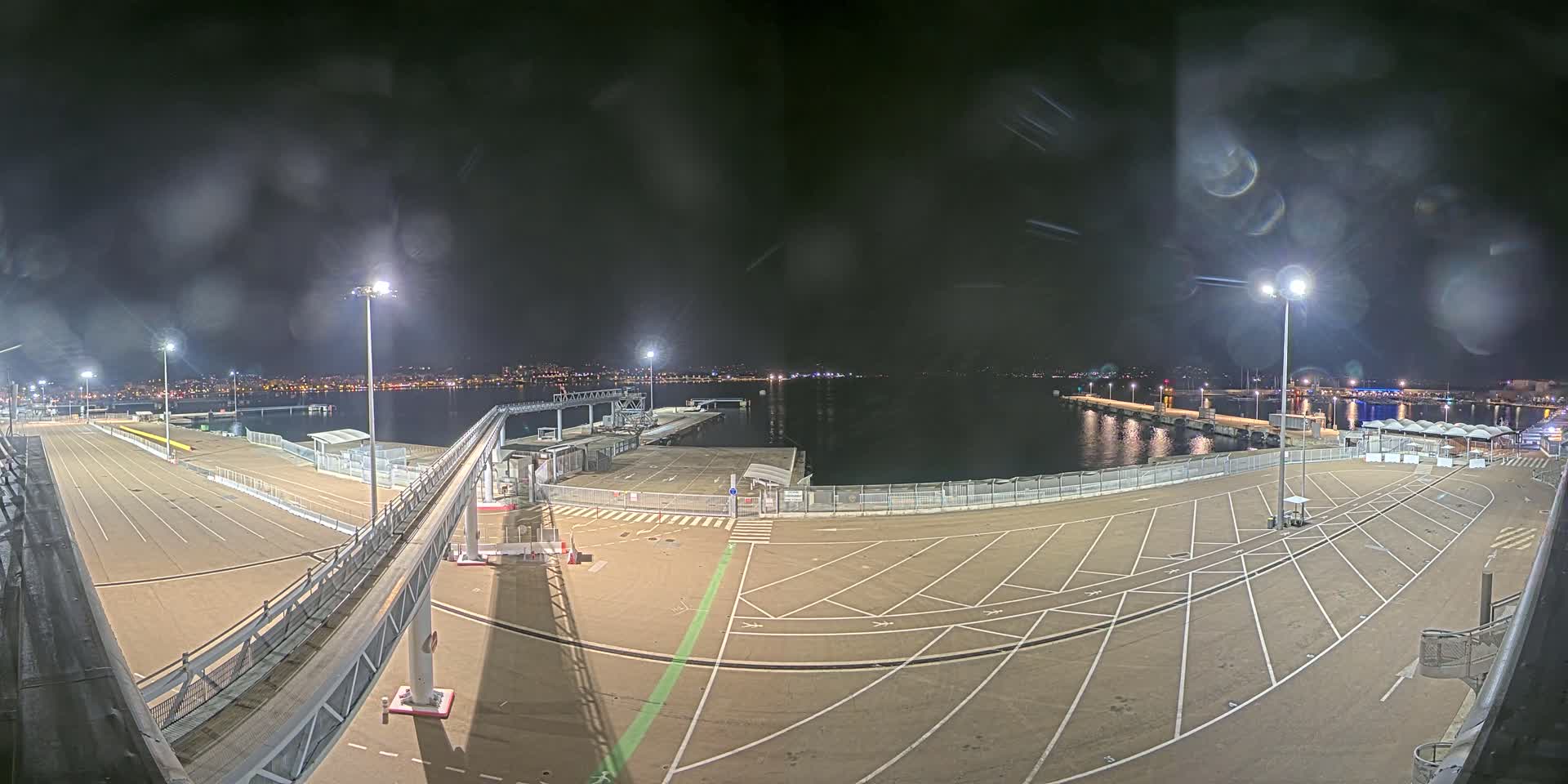 A nighttime view of an empty port area with a long gangway leading to a dock, illuminated by tall lampposts, overlooking a calm body of water and a city skyline in the distance.