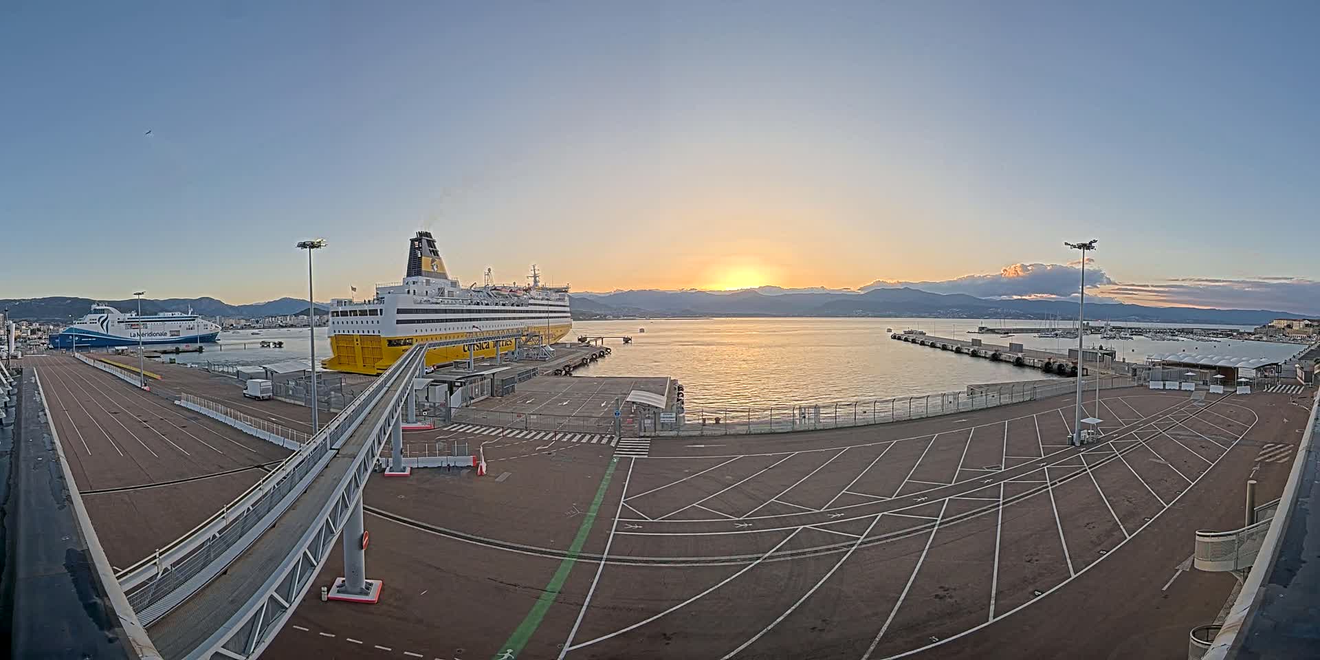 A large ferry is docked at a port during a calm sunset, with another smaller ferry visible in the distance.