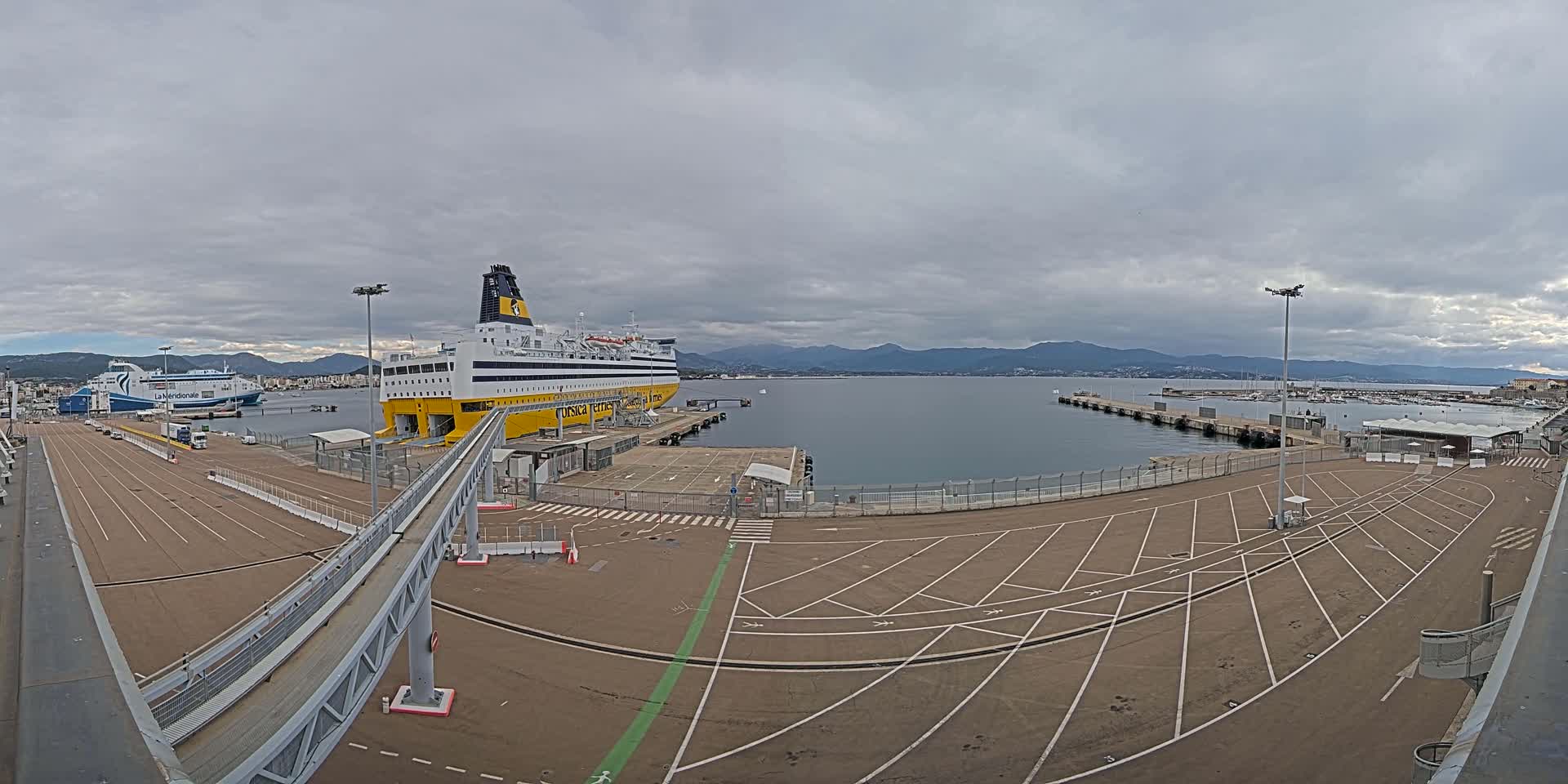 A large yellow and black ferry is docked at a port under an overcast sky, with another smaller ferry visible in the distance and mountains in the background.