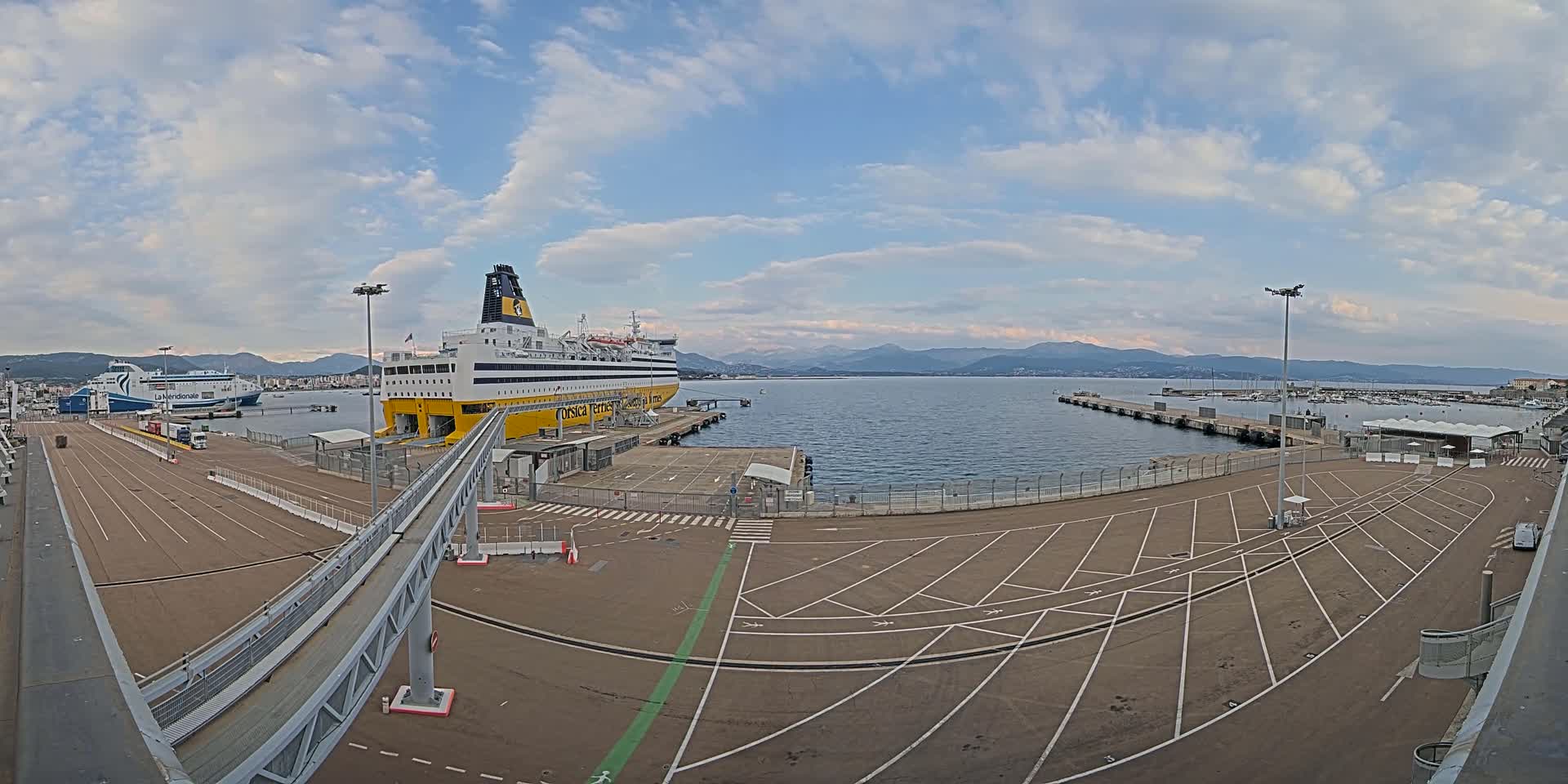 A large yellow and black ferry is docked at a port under a partly cloudy sky, with another ferry visible in the distance and mountains in the background.
