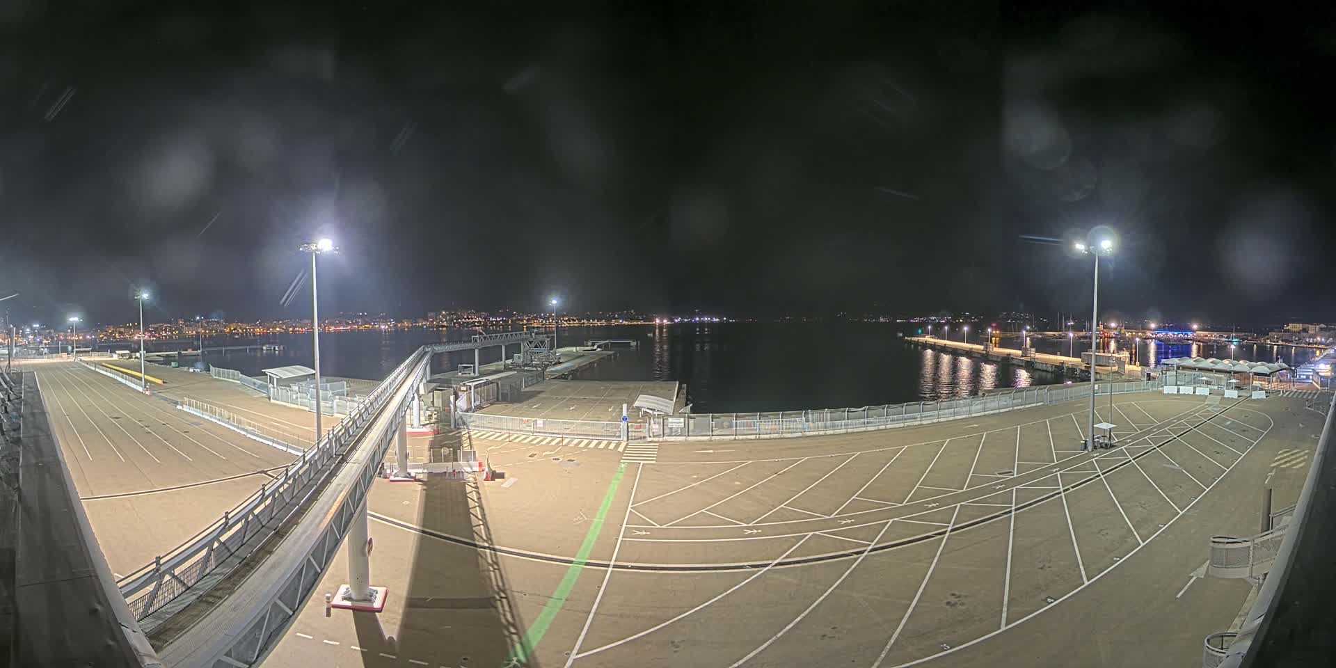 A nighttime view of an empty port area with a gangway, parking spaces, and a calm body of water in the background, under a slightly hazy sky.