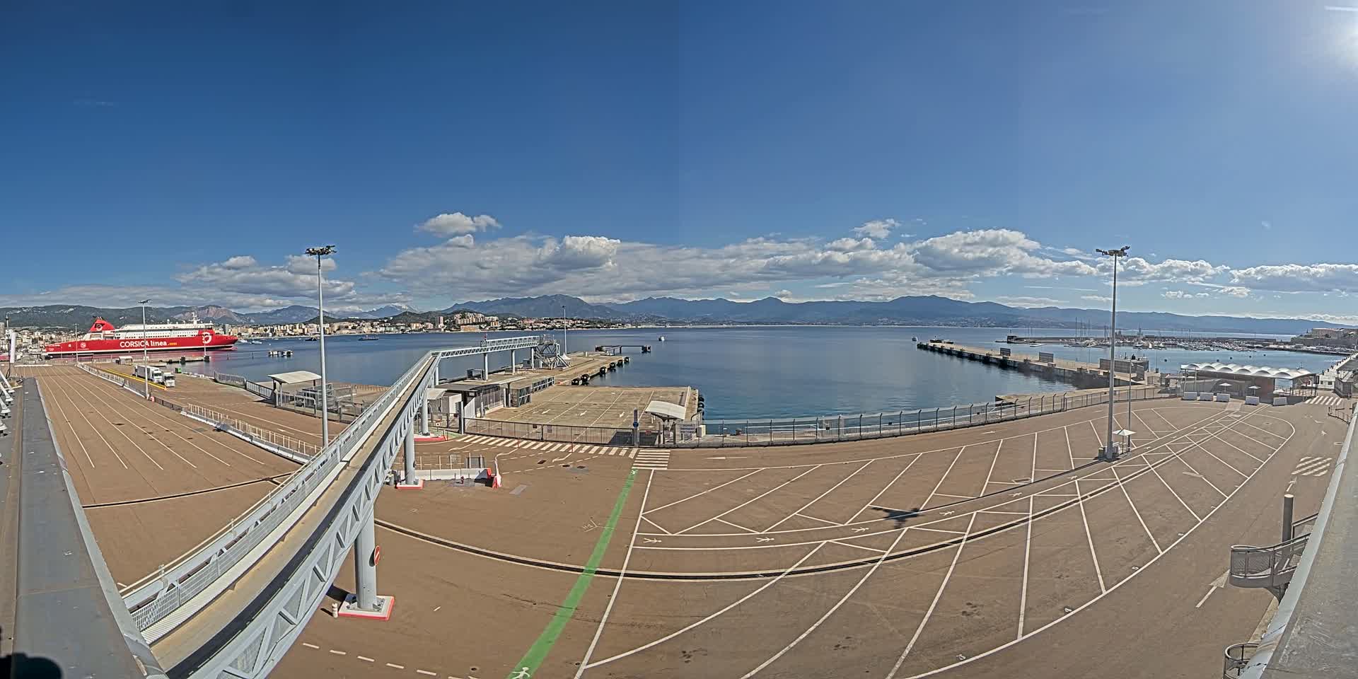 A large empty ferry terminal with a red ferry docked, under a mostly sunny sky with some clouds, overlooks a calm body of water and distant mountains.