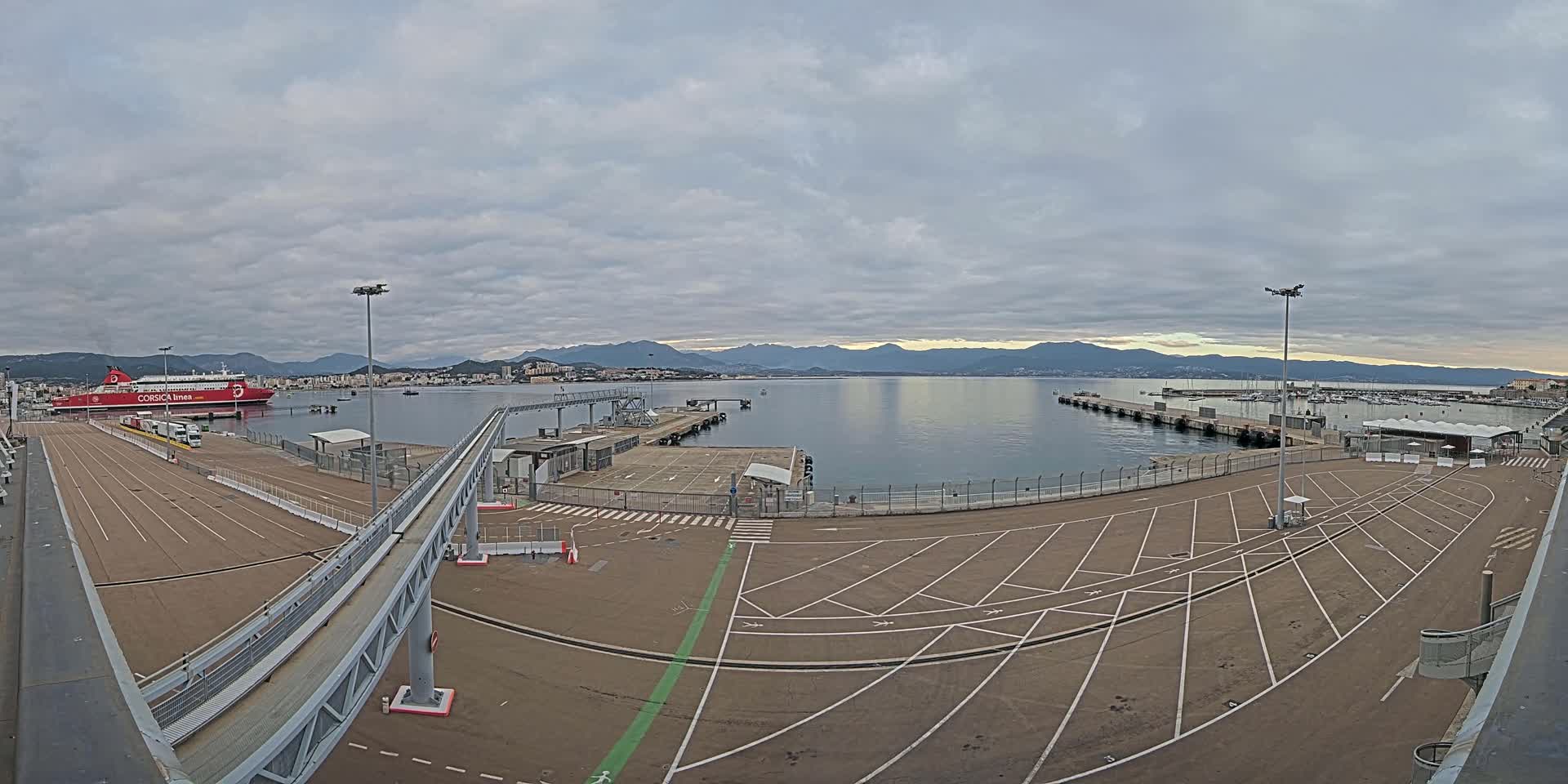 A large ferry is docked at a port, with an empty parking lot and a walkway leading to it, under a cloudy sky and mountains in the distance.