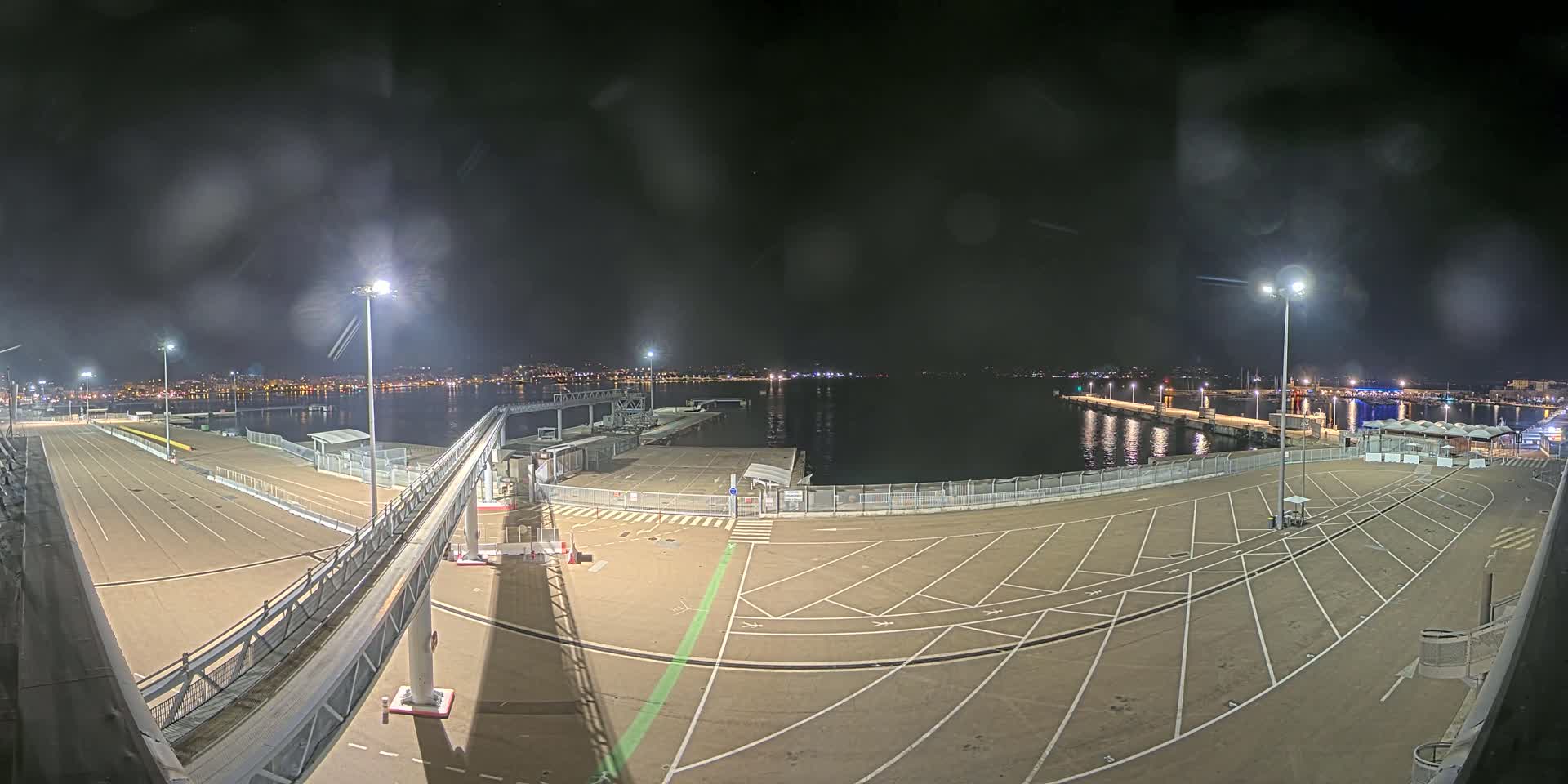 A nighttime view of an empty port area with marked parking spaces, a gangway, and a body of water in the background under a slightly cloudy sky.