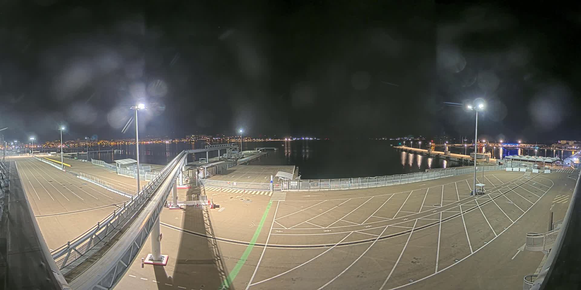 A nighttime view of an empty port area with a walkway and parking lot, illuminated by bright lights, under a slightly hazy sky.