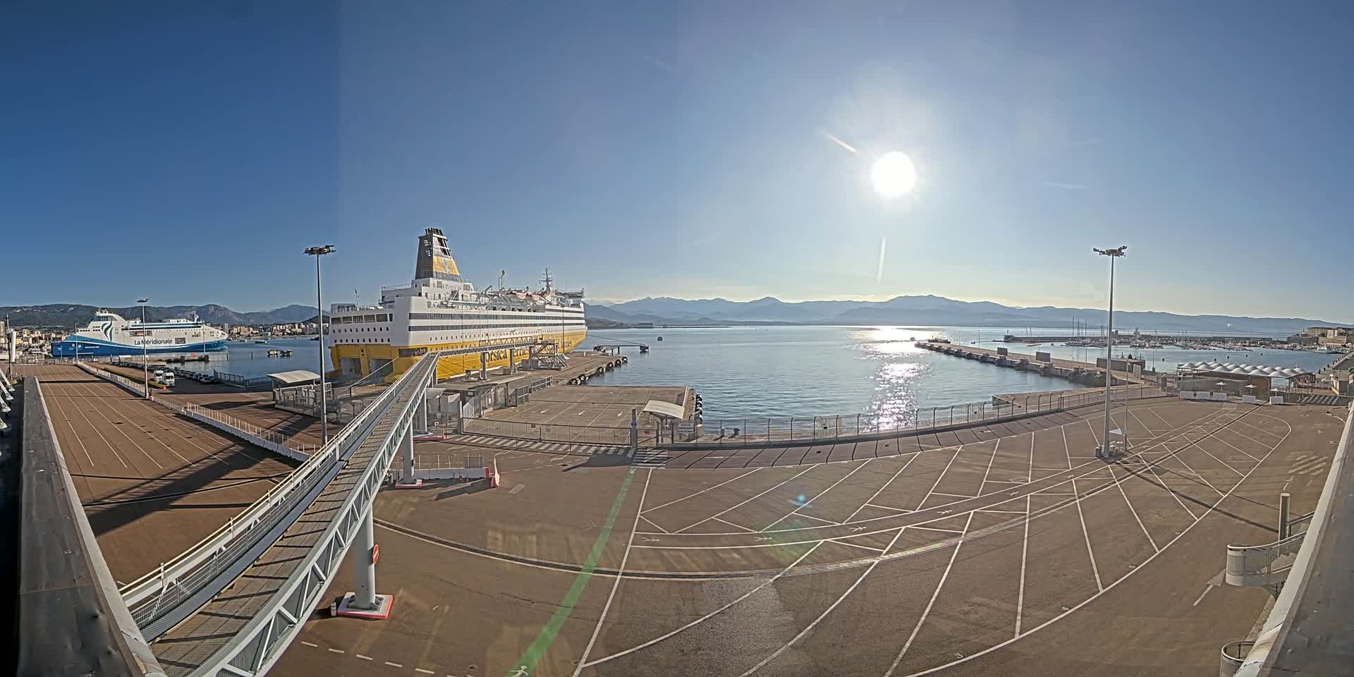 A large ferry is docked at a port on a sunny day, with another smaller ferry visible in the distance and mountains in the background.