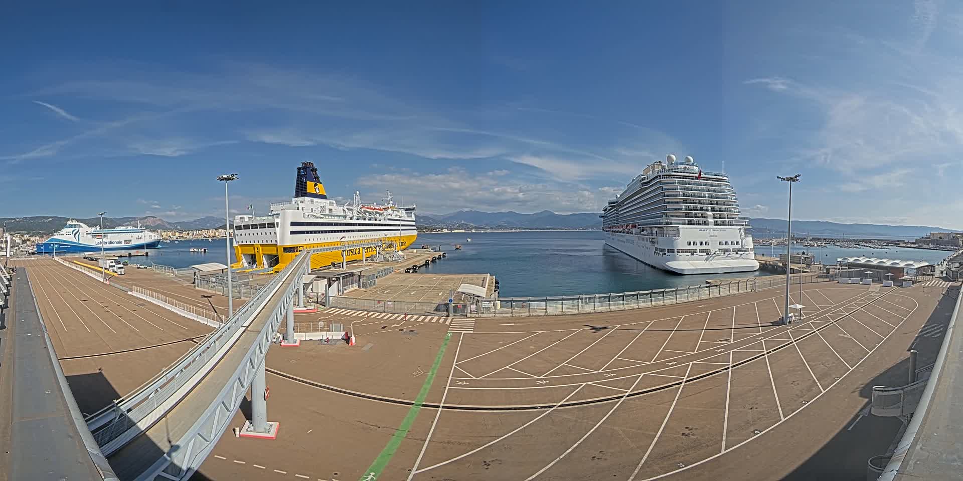 A large cruise ship and two ferries are docked at a port on a sunny day with mountains visible in the distance.