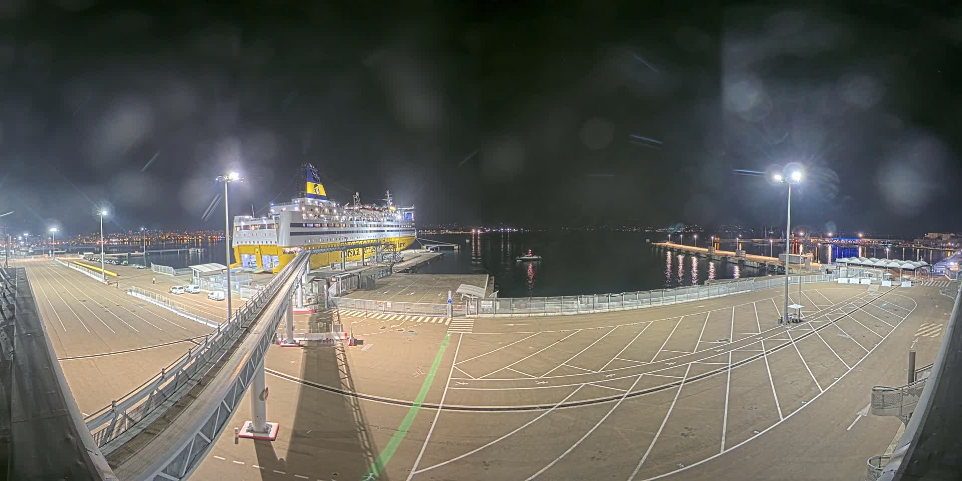 A large ferry is docked at a port at night under a slightly hazy sky, with a walkway connecting the ship to the terminal's empty parking lot.