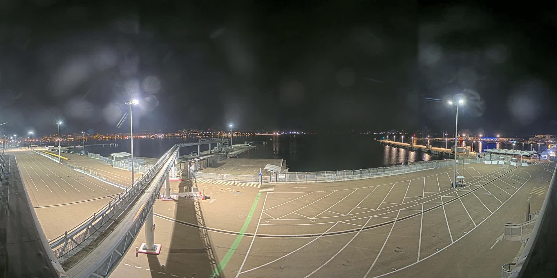 A nighttime view of a mostly empty port area with a gangway, parking spaces, and a body of water showing a distant city skyline under a dark, slightly rainy sky.