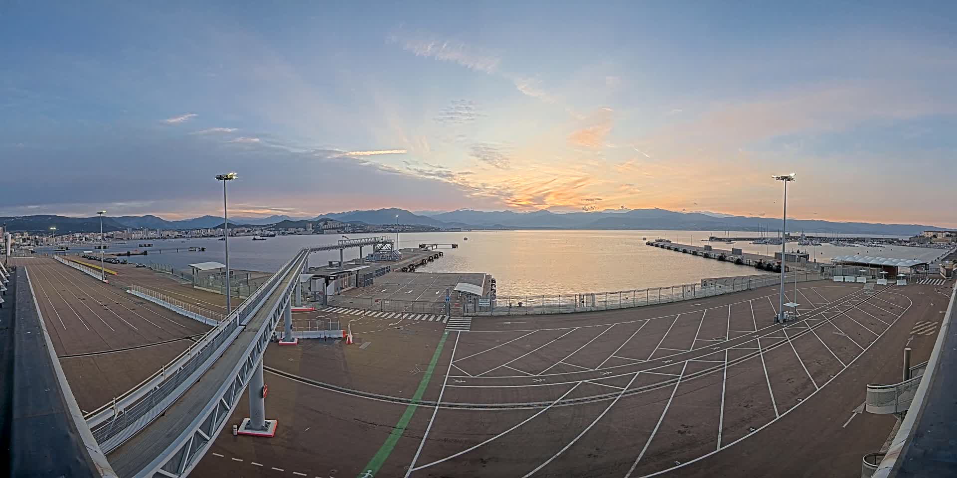 An empty harbor parking lot at sunset overlooks a calm body of water with mountains in the background.