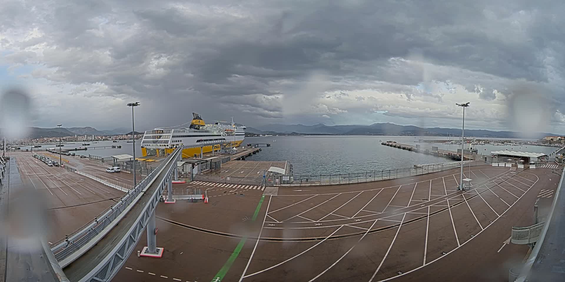 A large ferry is docked at a port under a cloudy, possibly rainy sky, with a long walkway leading to it and an empty parking lot in the foreground.