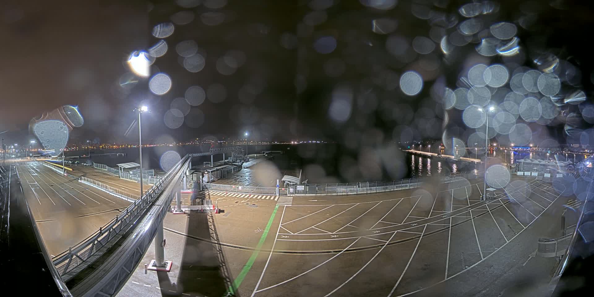 A nighttime view of a ferry terminal's empty parking lot and gangway, seen through rain-covered glass, with city lights visible in the distance.