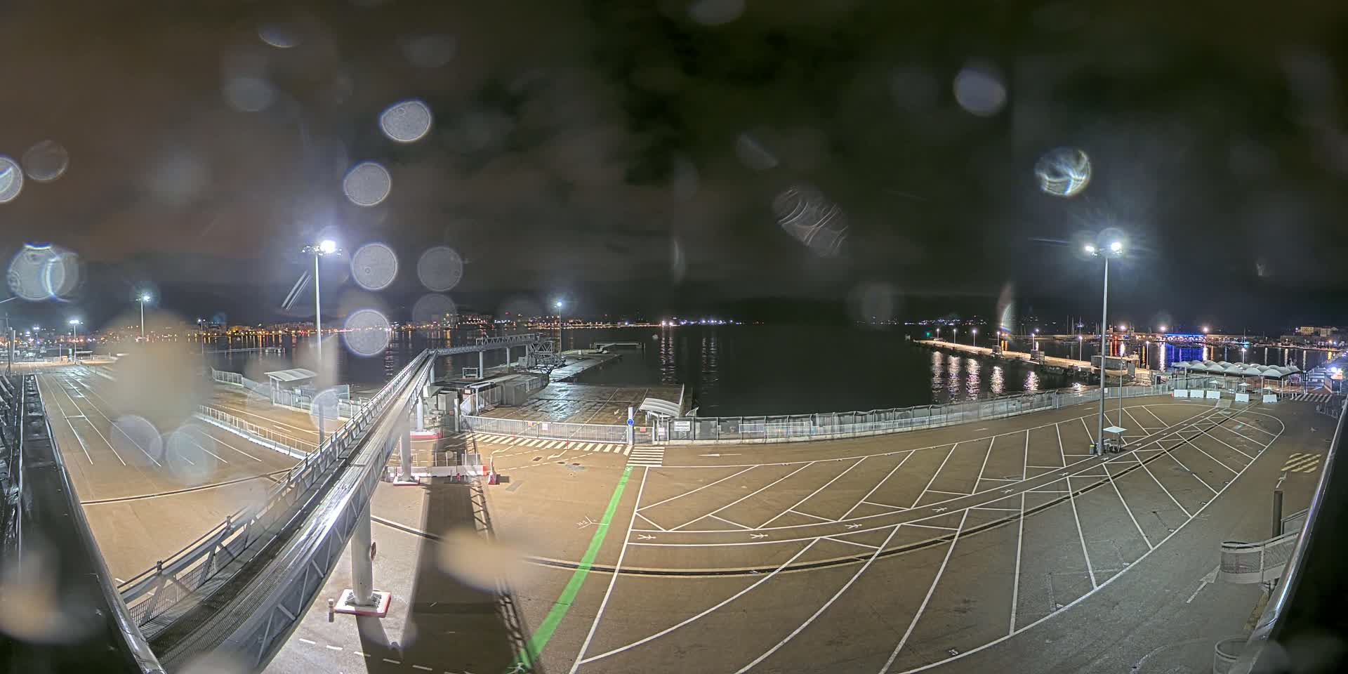 A nighttime view of a rain-soaked port area with empty parking lots, a gangway, and a calm body of water reflecting city lights in the distance.