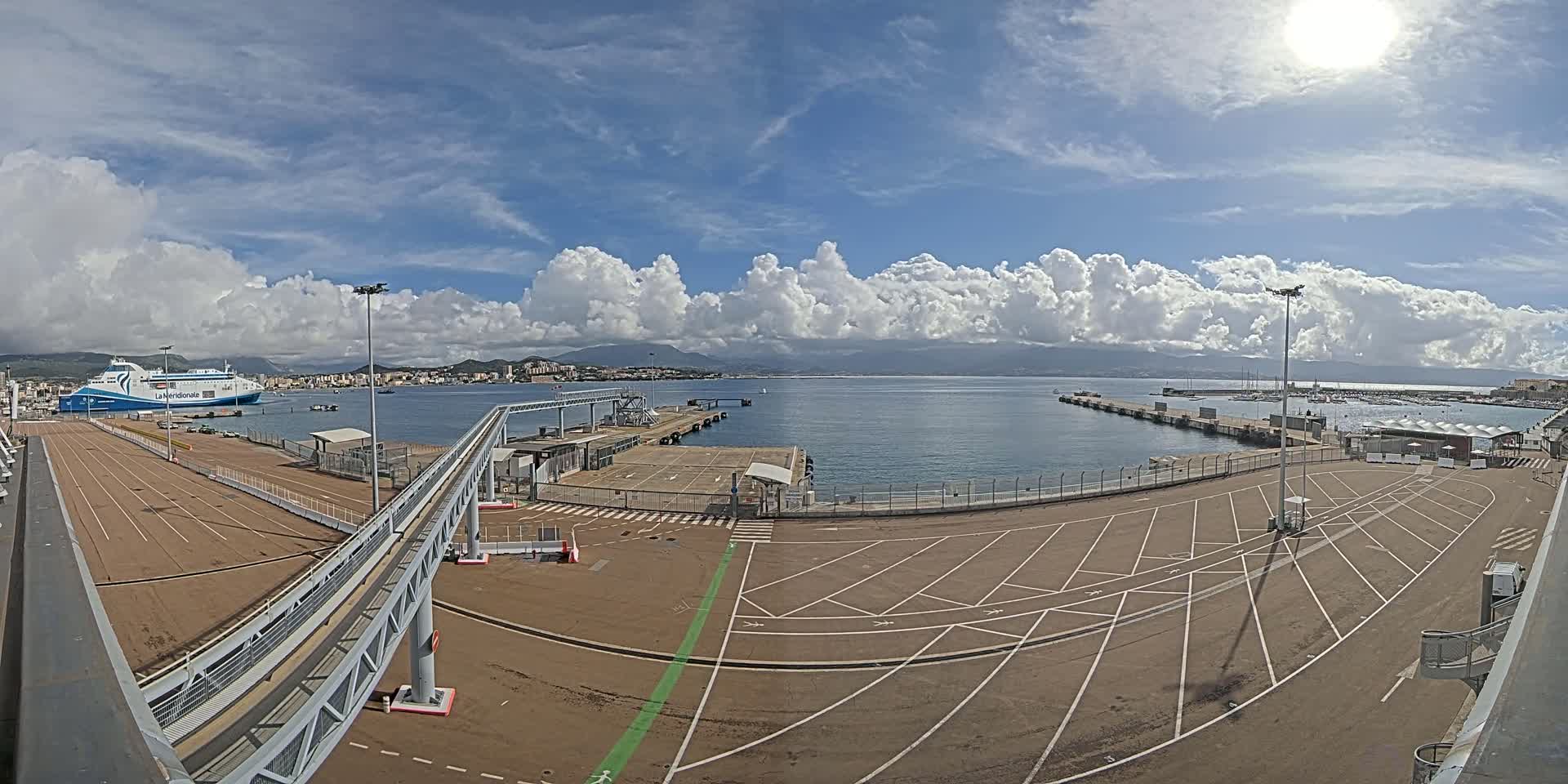 A ferry is docked at a port under a partly cloudy sky, with a large empty parking lot and a walkway visible in the foreground.