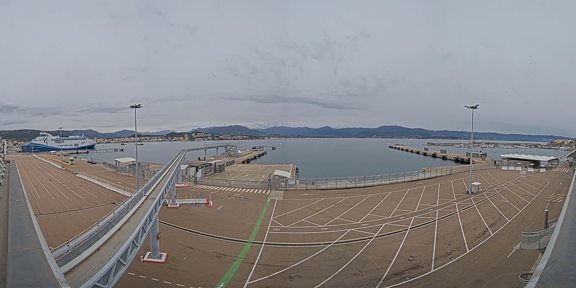 A ferry is docked at a port with a large empty parking lot, under an overcast sky, and mountains visible in the distance.