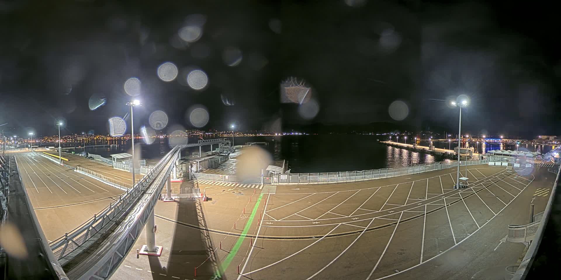 A nighttime view of a ferry terminal with rain-streaked glass obscuring the view, showing an empty parking lot, gangway, and calm water beyond.
