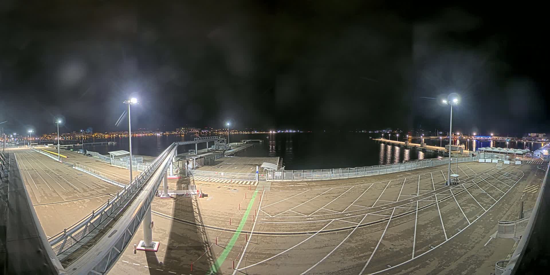 An empty, nighttime port area with a gangway leading to a dock, illuminated by several lights, overlooks a calm body of water and a distant city skyline.