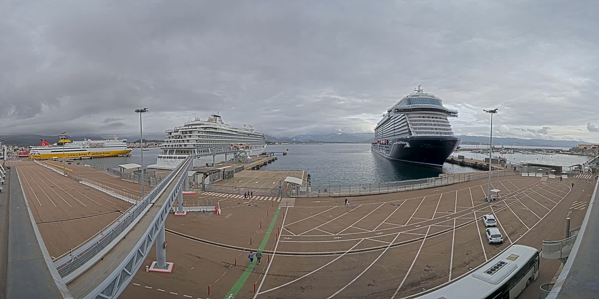 A cruise ship and a ferry are docked at a port under a cloudy sky, with a parking lot and pedestrian walkways visible.