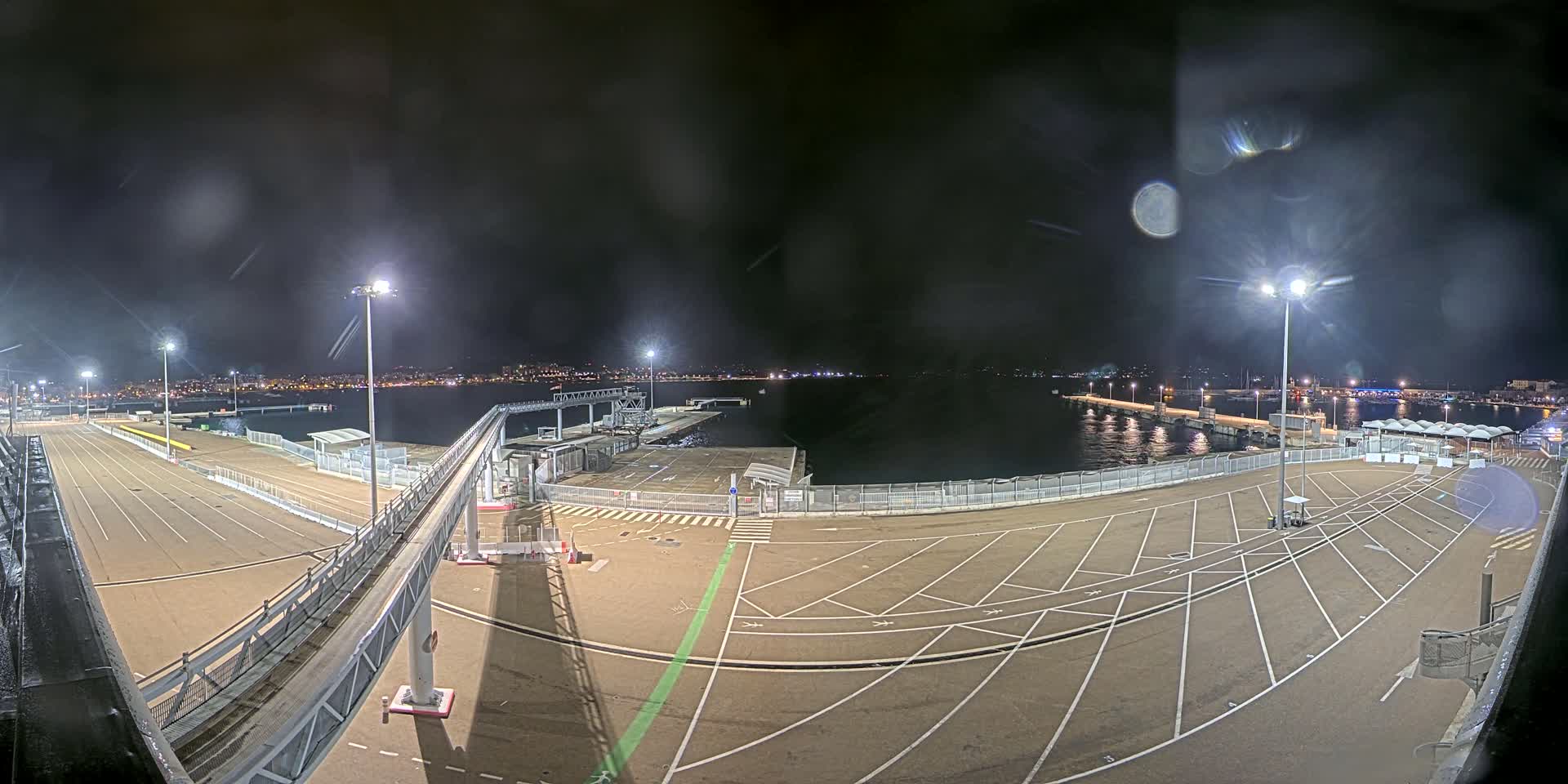 An empty, well-lit nighttime port area with a gangway leading to a dock, overlooking a calm body of water and a city skyline in the distance.