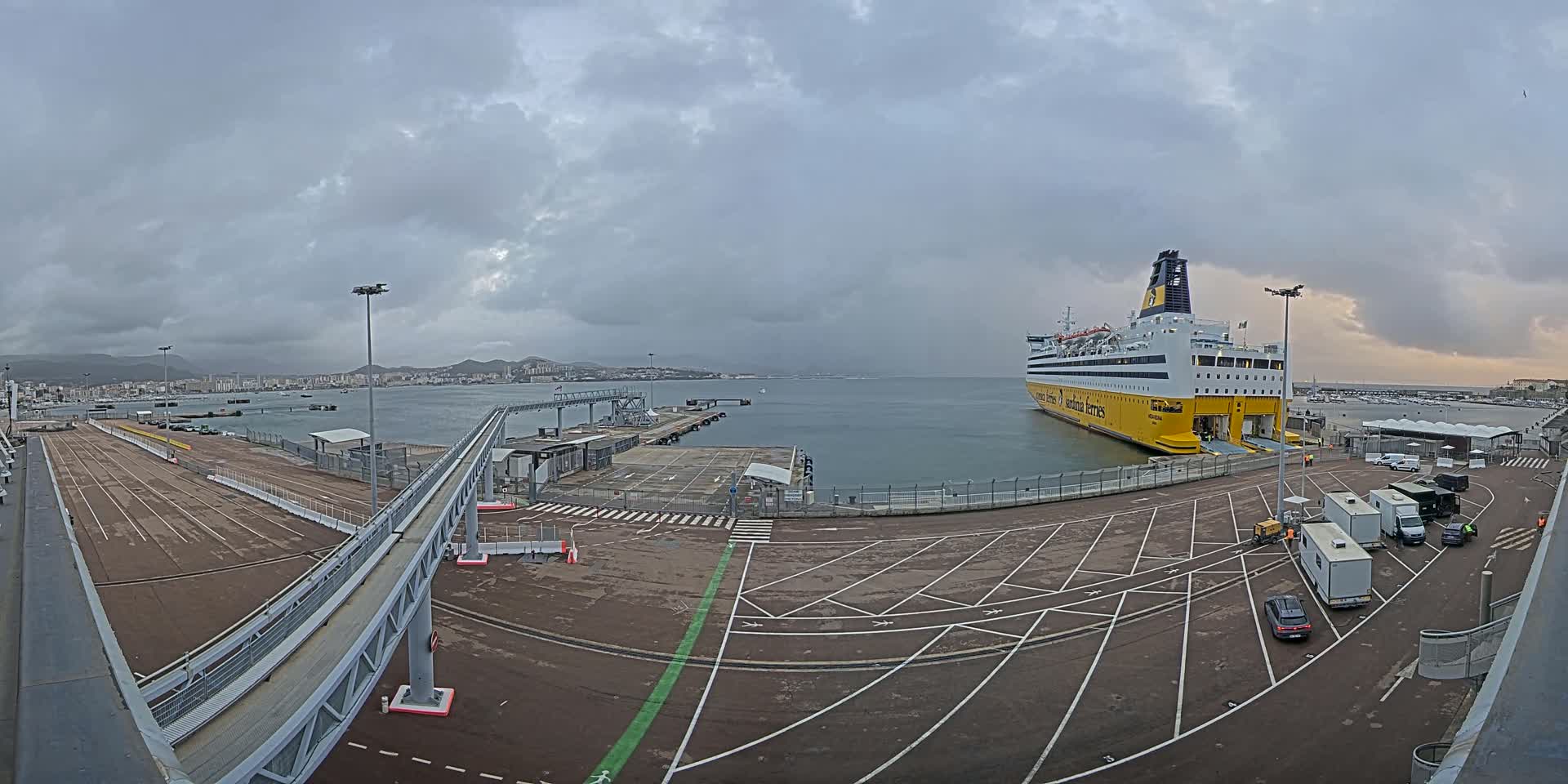 A large yellow and white ferry is docked at a port under an overcast sky, with a paved area and several vehicles visible.