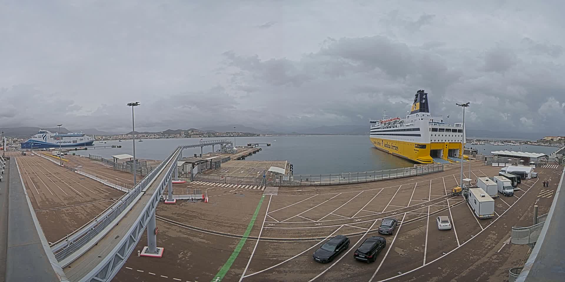 A large ferry is docked at a port under an overcast sky, with several cars parked in the lot and another ferry visible in the distance.